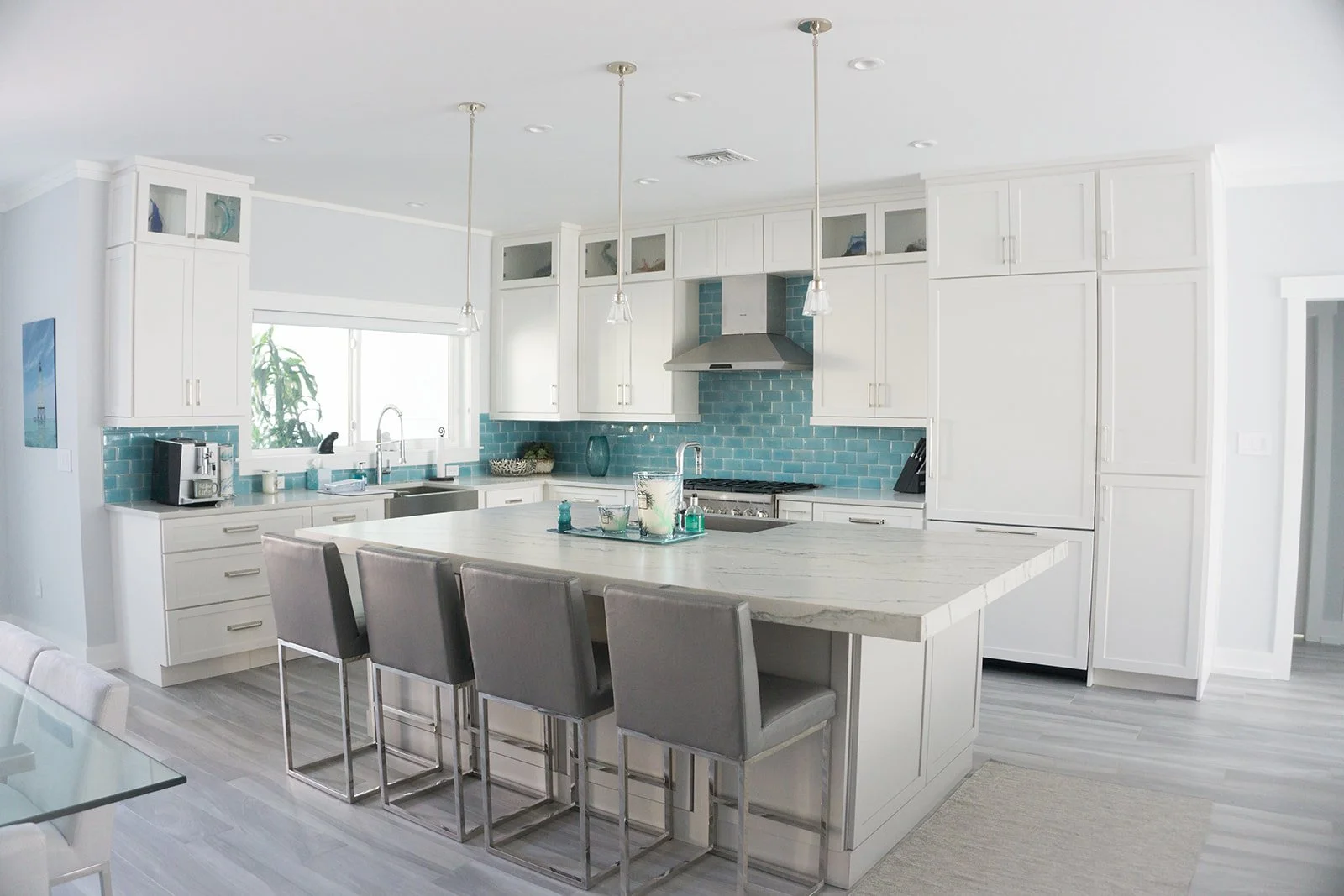 Modern kitchen with white cabinets, turquoise subway tile backsplash, kitchen island with four barstools, stainless steel range hood, and window above sink.