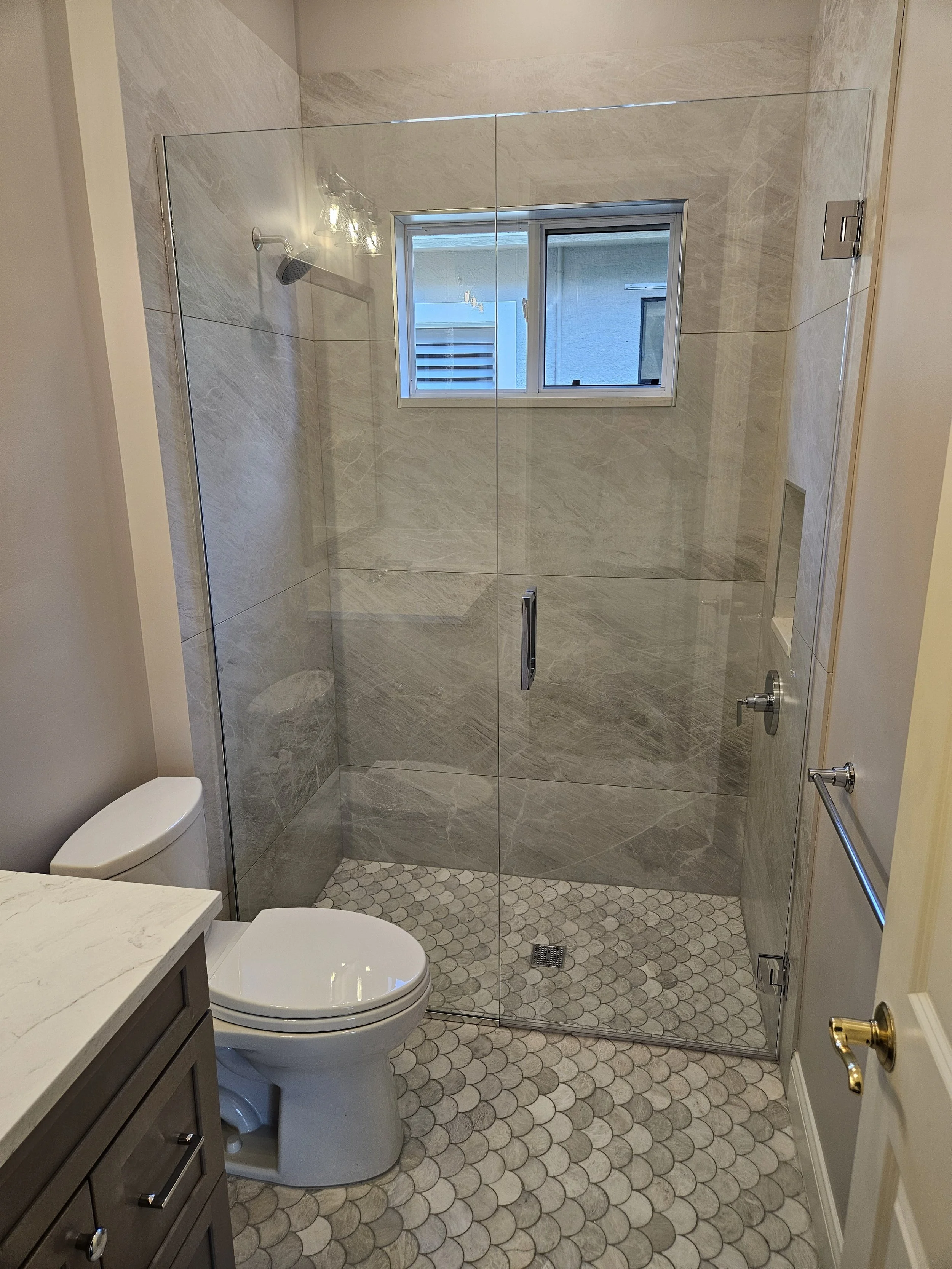 Bathroom with shower area enclosed by glass door, toilet, and a small window, with beige tiled walls and hexagonal floor tiles.