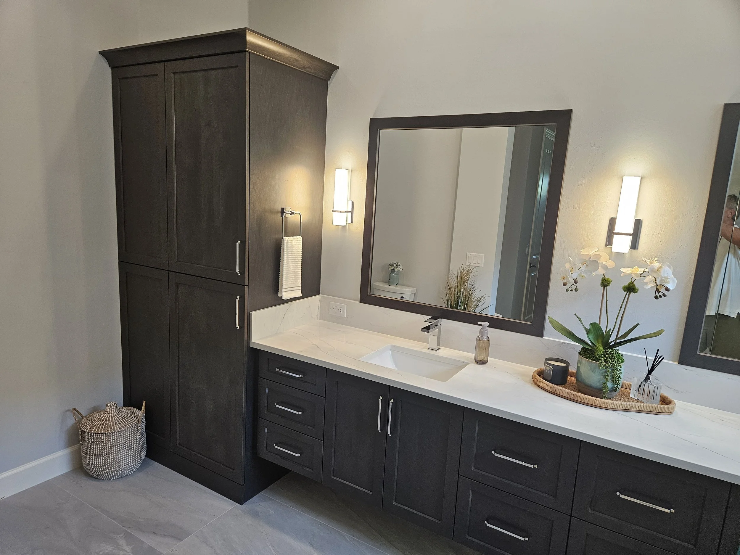 Modern bathroom vanity with dark cabinets, white countertop, and rectangular mirror. Decor includes orchids and plants on the counter, with wall-mounted lights and a towel hanging on the wall.