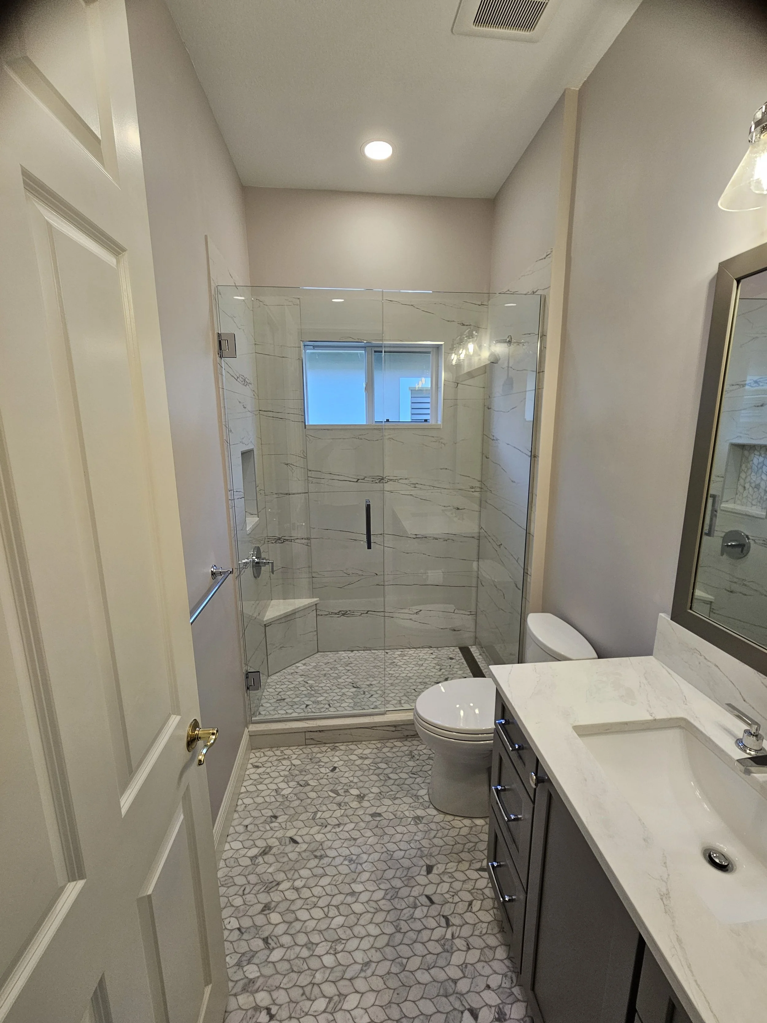 Interior view of a modern bathroom with a glass shower enclosure, a white toilet, a marble countertop with a sink, a mirror, and patterned tile flooring.