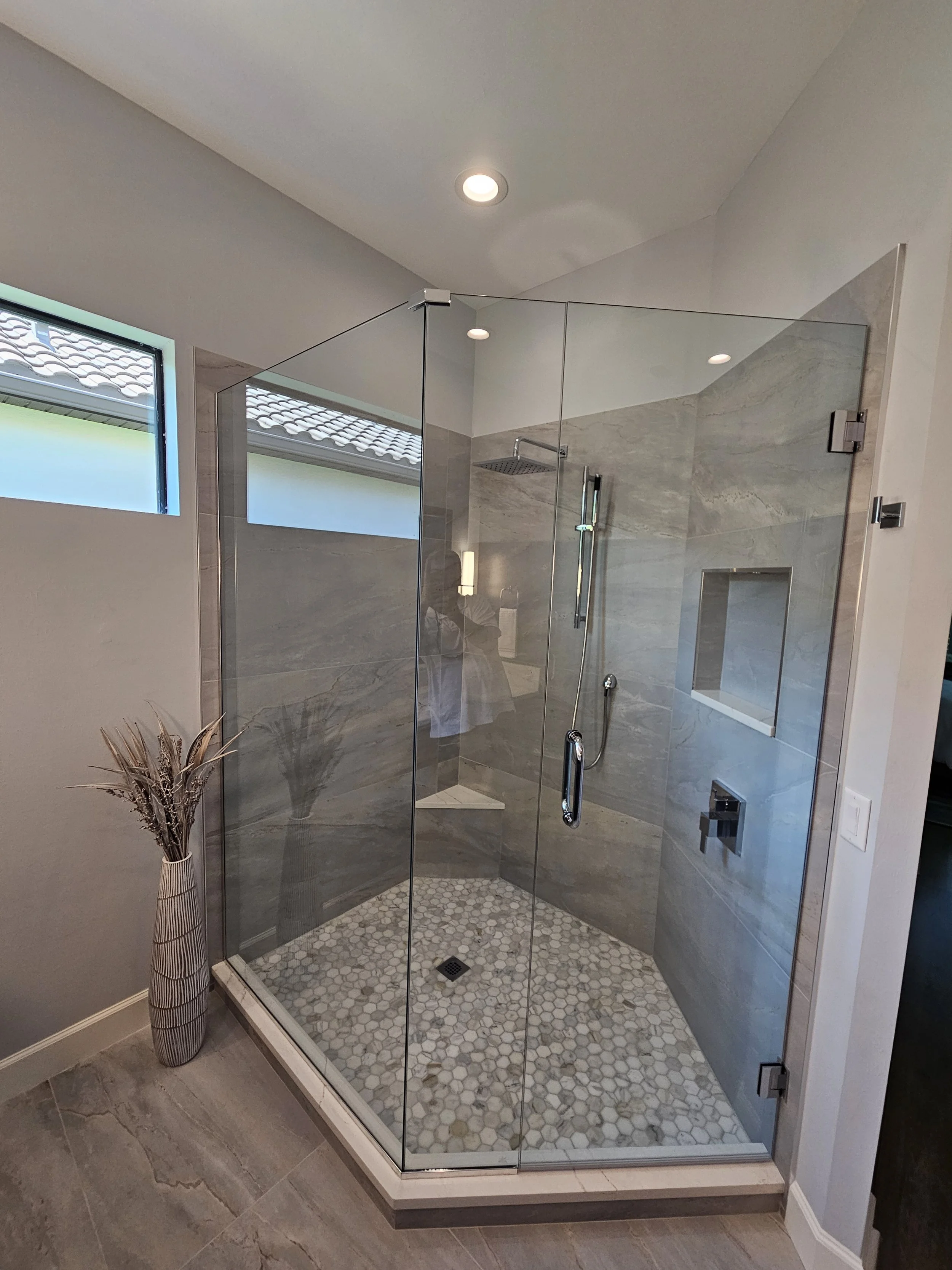 A modern walk-in shower with glass enclosure, gray marble tiles on the walls, hexagonal pattern tiles on the floor, a showerhead, and a built-in niche for toiletries. There is a window with frosted glass and a decorative floor vase with dried plants 
