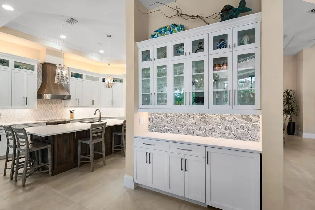 Kitchen with white cabinets, a kitchen island with a white countertop, and a built-in glass-front cabinet with decorative items and glassware. Pendant and ceiling lights illuminate the area.
