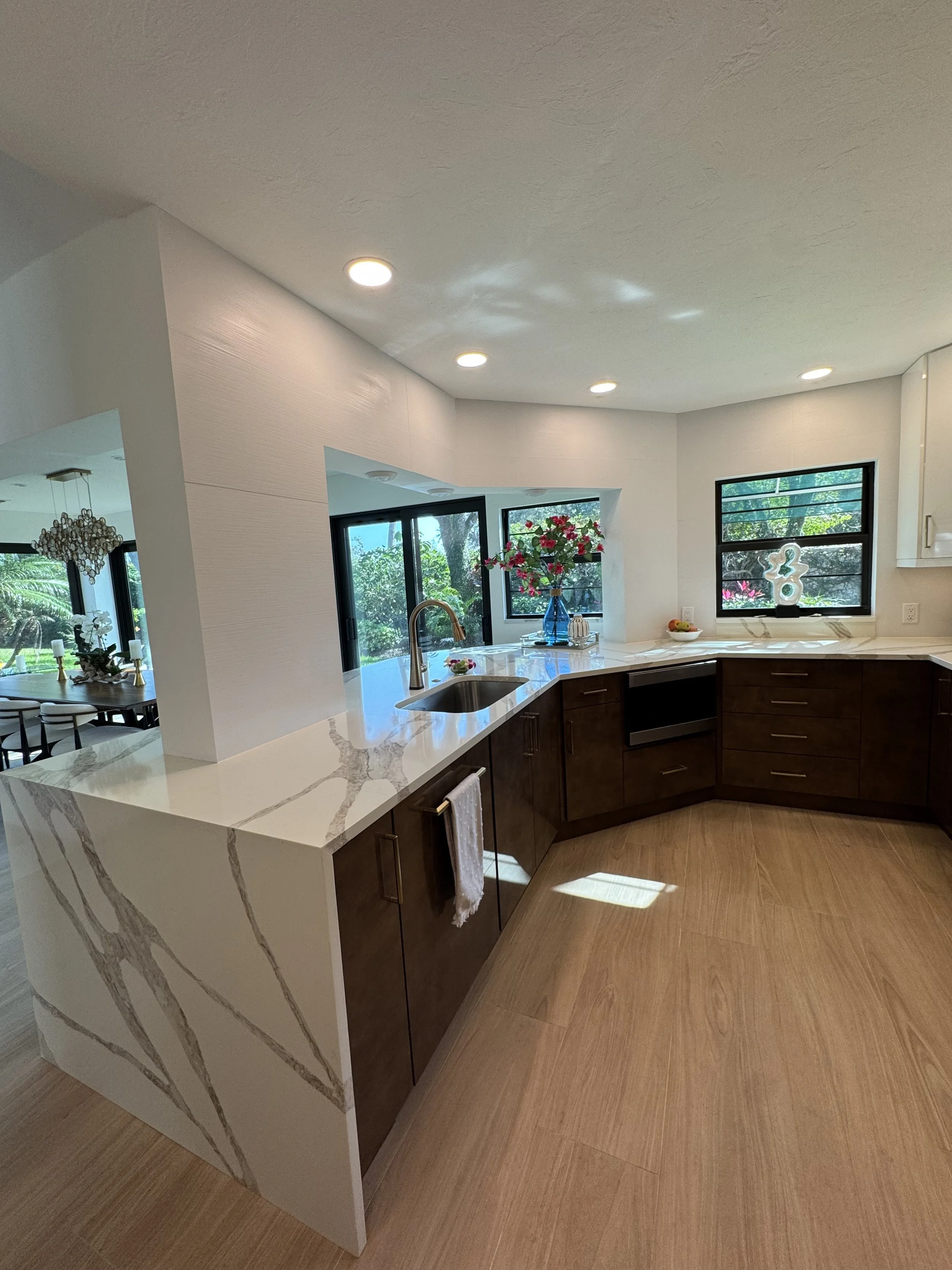 Modern kitchen with white marble countertop, brown cabinets, a sink, and windows overlooking a green outdoor area.
