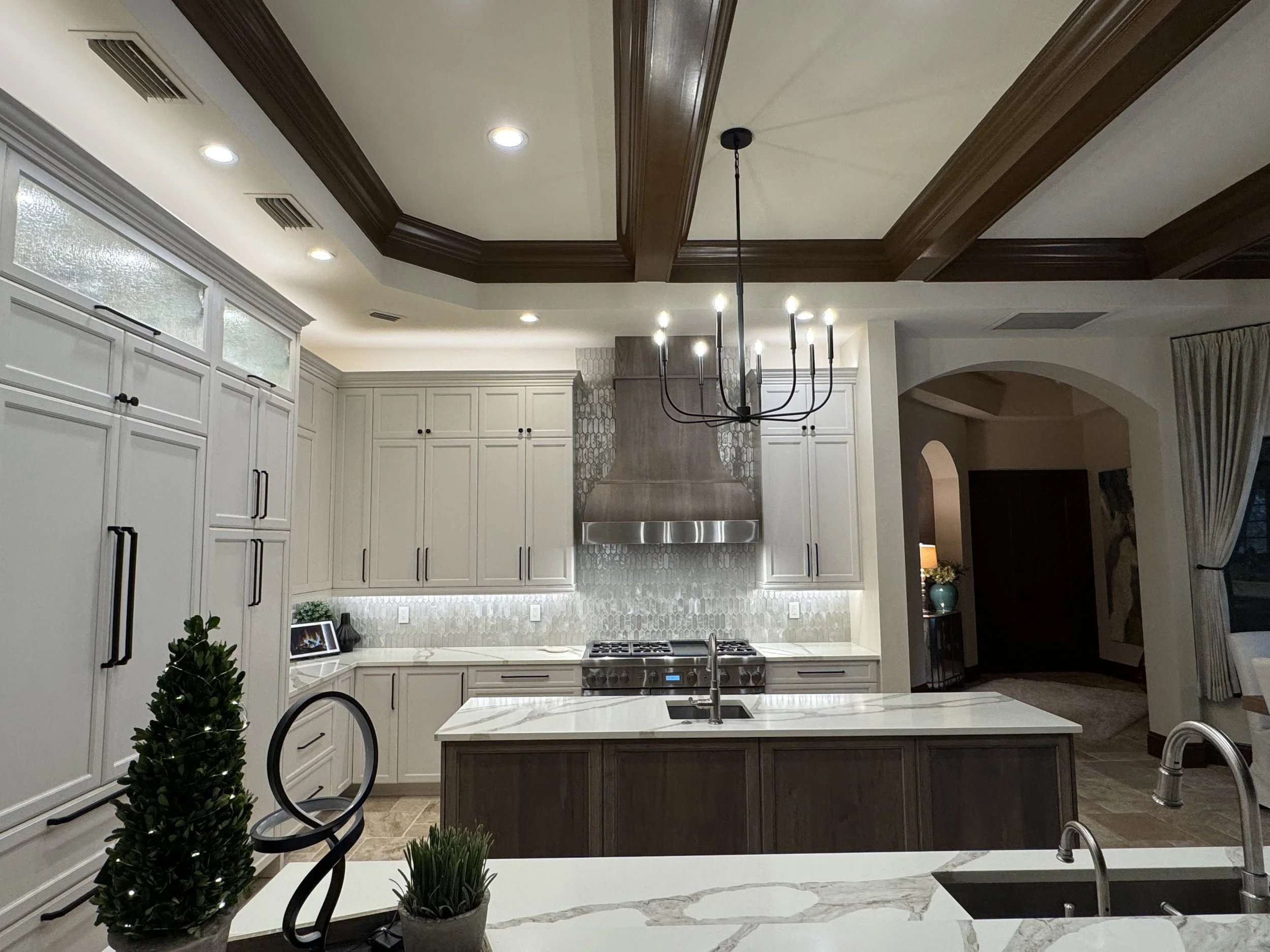 Modern kitchen with white cabinets, marble island, stainless steel stove, and hanging black chandelier, with ceiling lights and a small Christmas tree in the foreground.