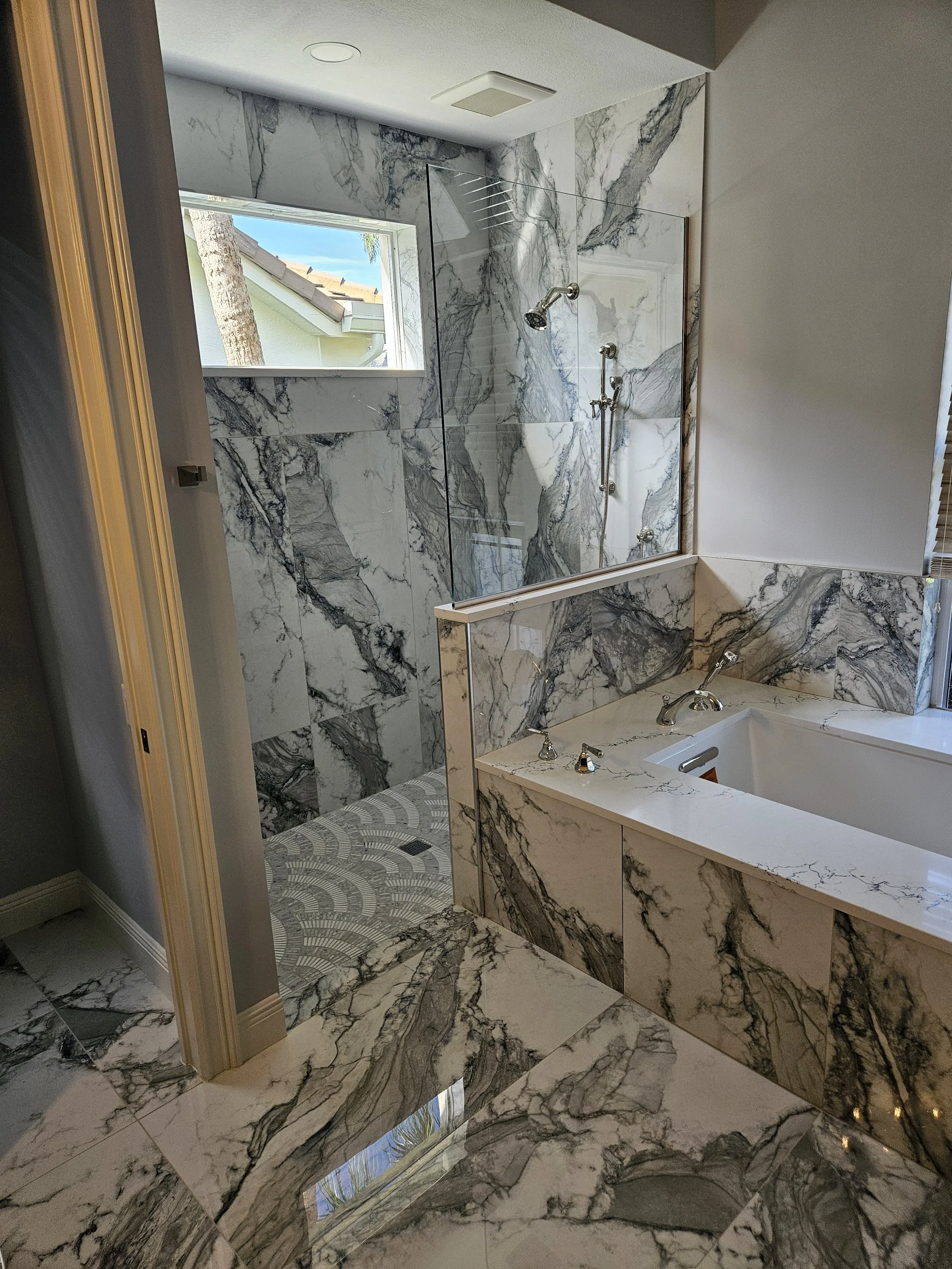 A luxurious bathroom with marble walls and floor, a walk-in shower with a glass partition, and a small window showing part of a tree and blue sky.