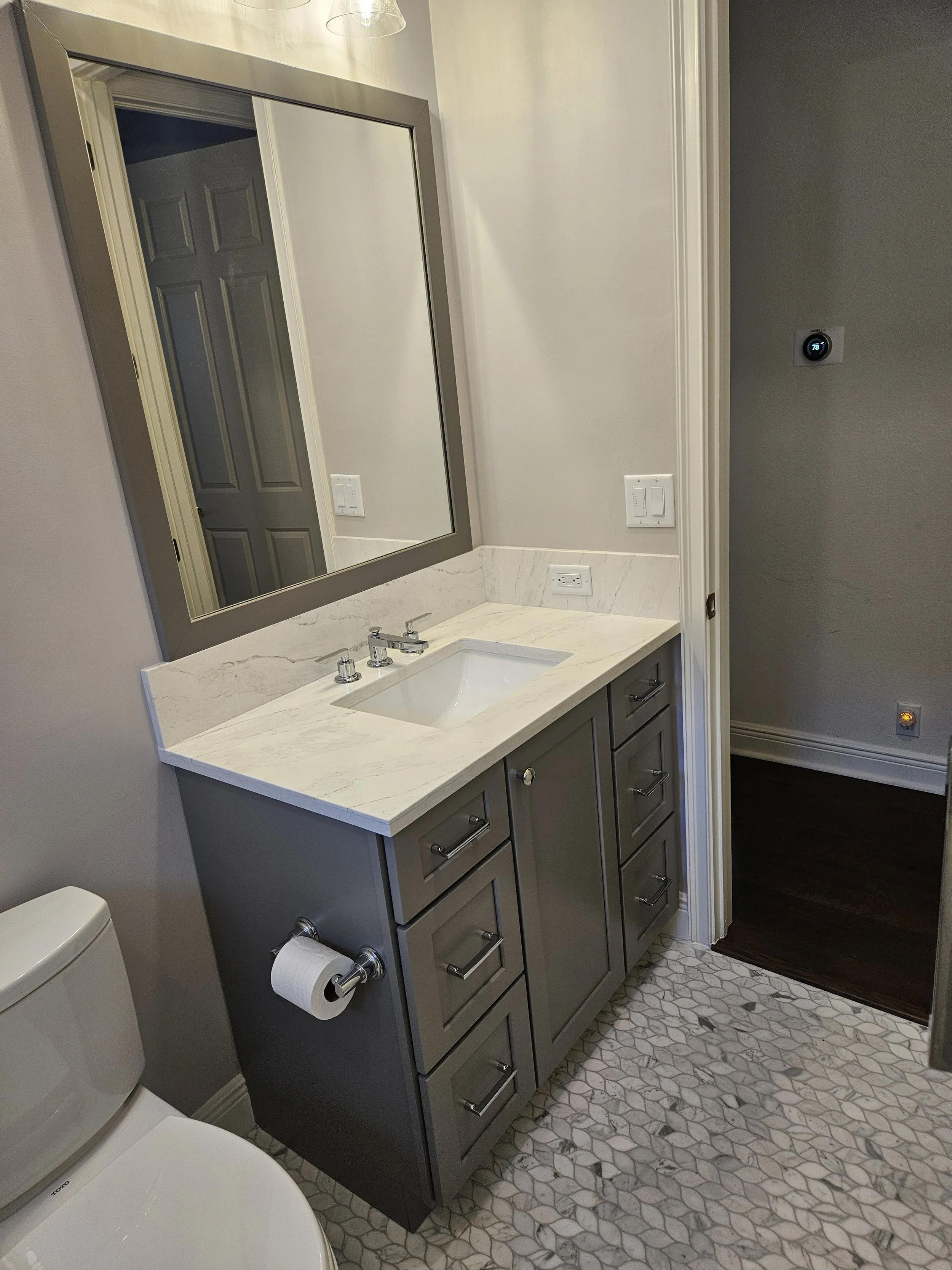 A bathroom vanity with a large mirror, a marble countertop, a white sink, and gray cabinets. To the left, a toilet paper holder with a roll of toilet paper, and a toilet below. The wall has a thermostat and a light switch. The floor has a hexagonal t