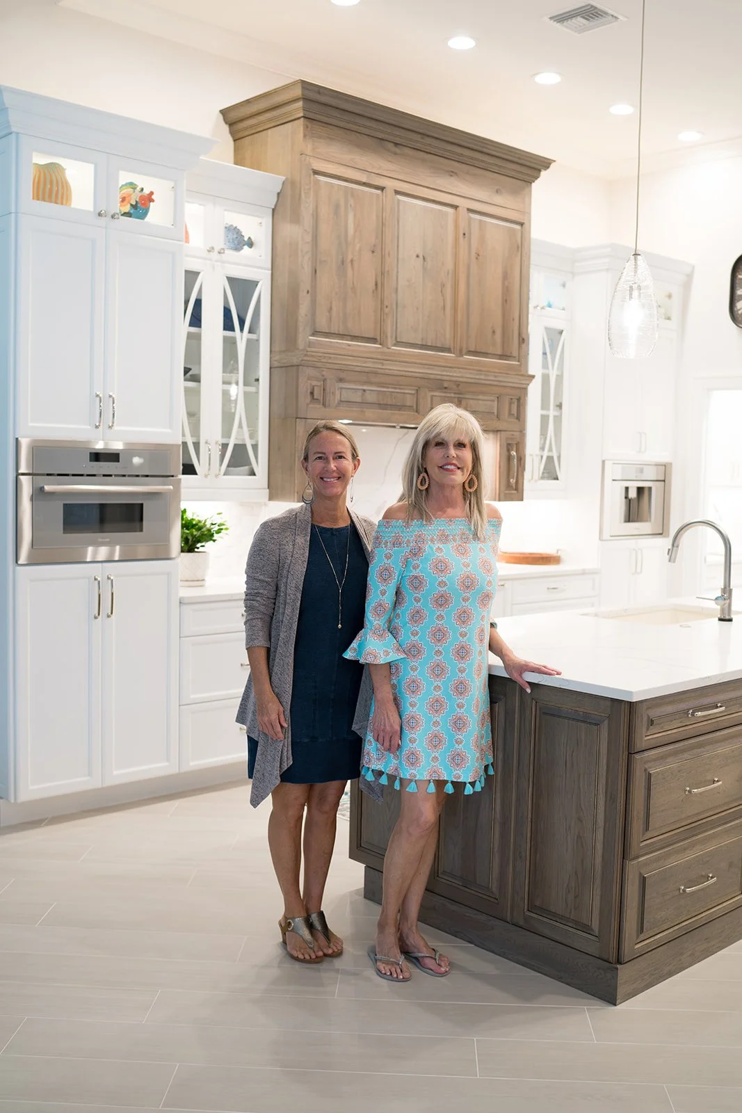 Two women standing in a modern kitchen with white and wood cabinetry, smiling at the camera.