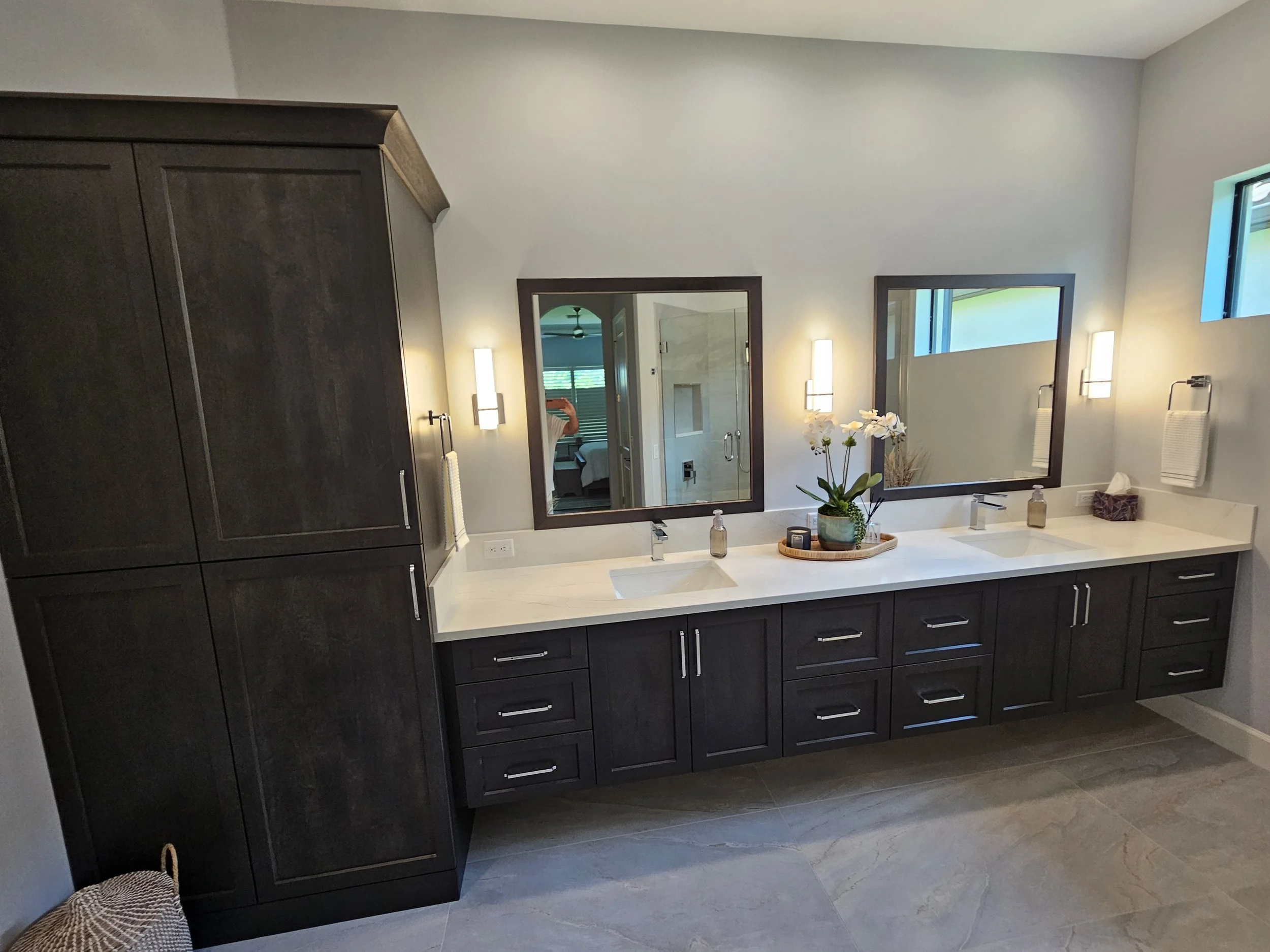 Bathroom with two mirrors, dark vanity cabinets, white countertop, and two sinks. Decor includes a potted orchid and toiletries. There are wall-mounted lights and window with natural light, with a view of a nearby bedroom.