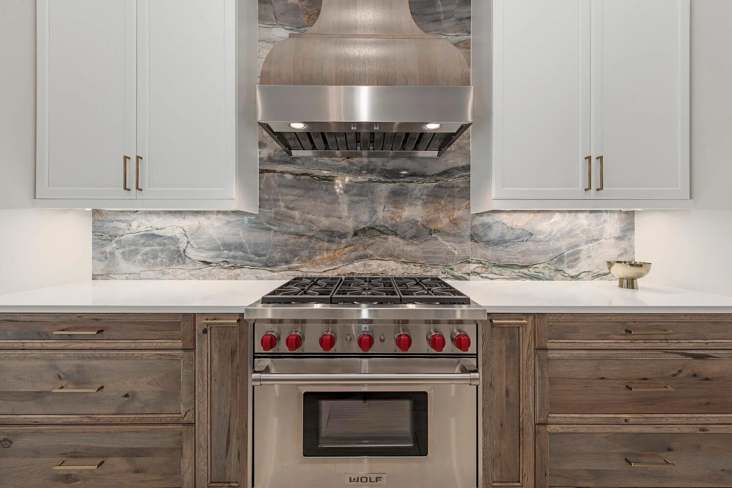 Modern kitchen with stainless steel Wolf oven, gray cabinets with gold handles, gray and beige marble backsplash, white countertops, and a decorative bowl on the right.