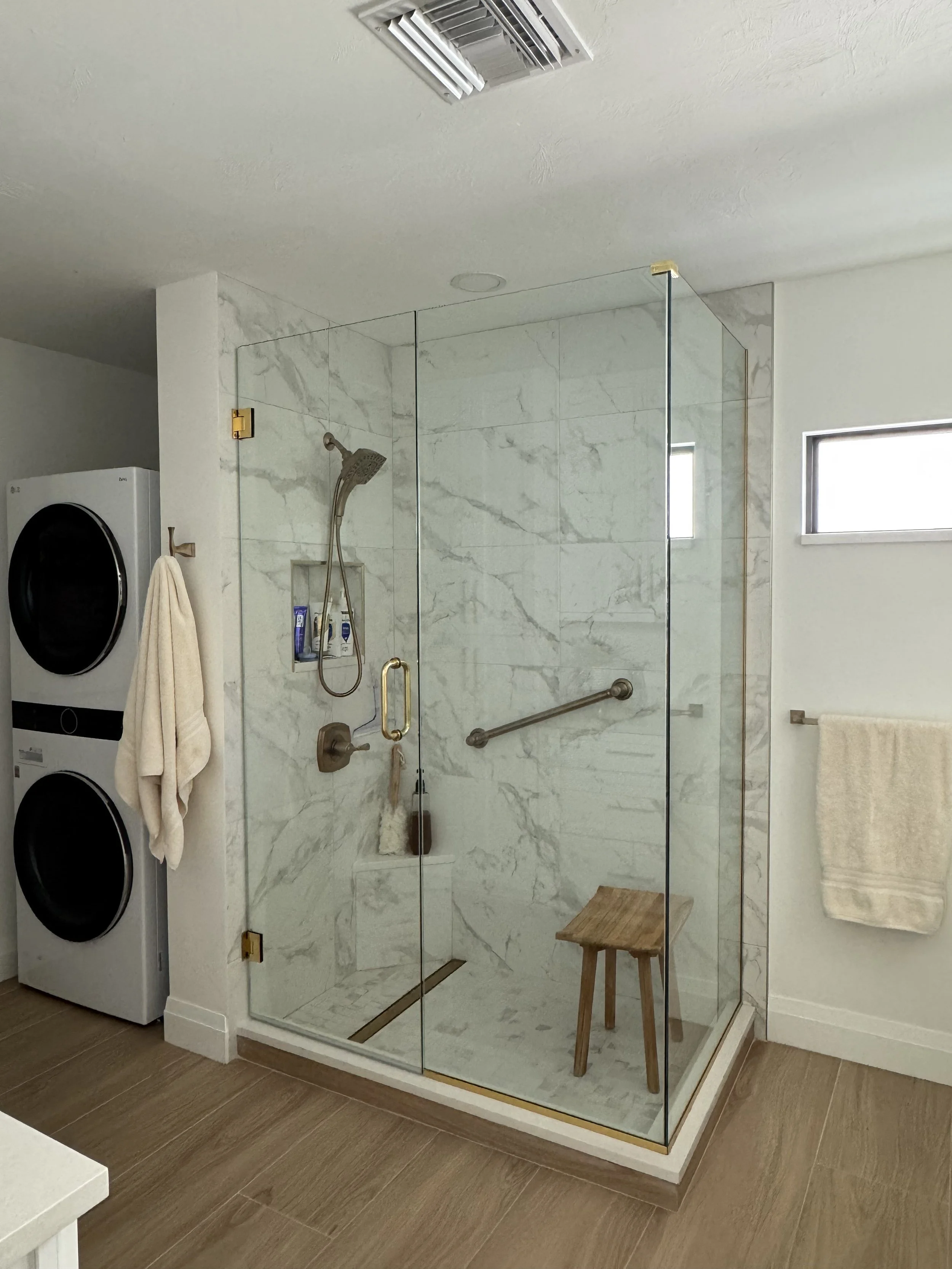 A modern bathroom with a glass-enclosed shower featuring marble walls, a rainfall showerhead, and a small wooden stool inside. To the left, there is a stacked washer and dryer, with a towel hanging on a hook. The floor has large brown tiles, and a to
