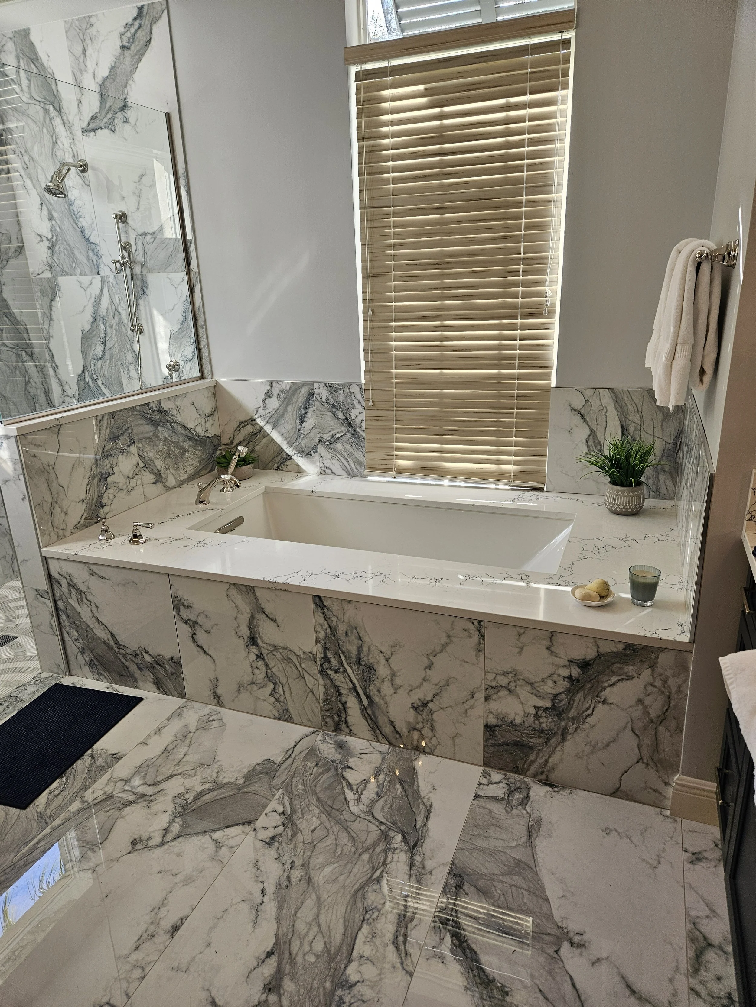 A luxurious bathroom with a marble bathtub, marble walls, and a large window with wooden blinds. There are two small plants, a towel rack with a white towel, and a tray with decor stones, a candle, and a small bottle.