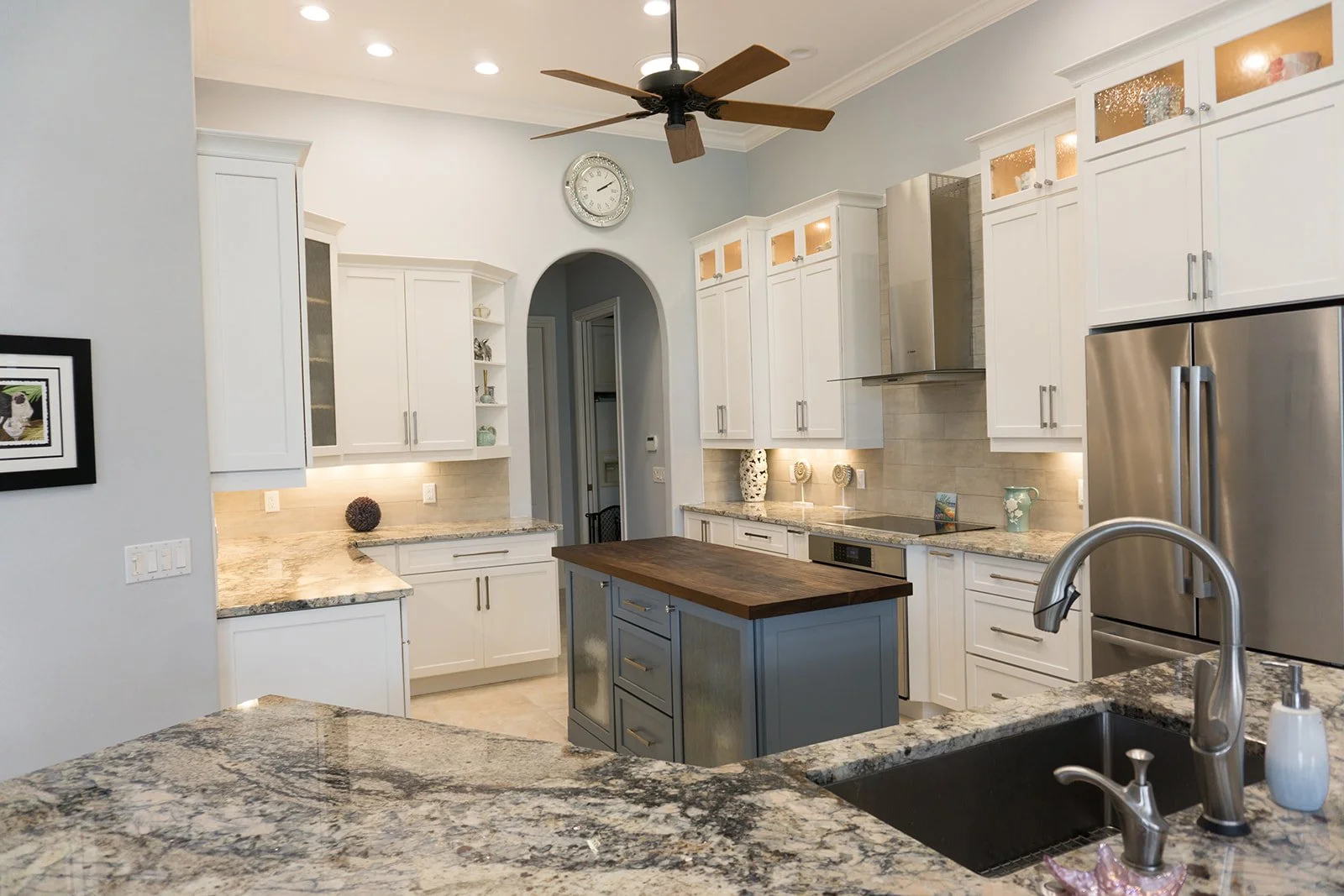Modern kitchen with white cabinets, granite countertops, stainless steel refrigerator, central island with wooden top, ceiling fan, and decorative vases on the counters.