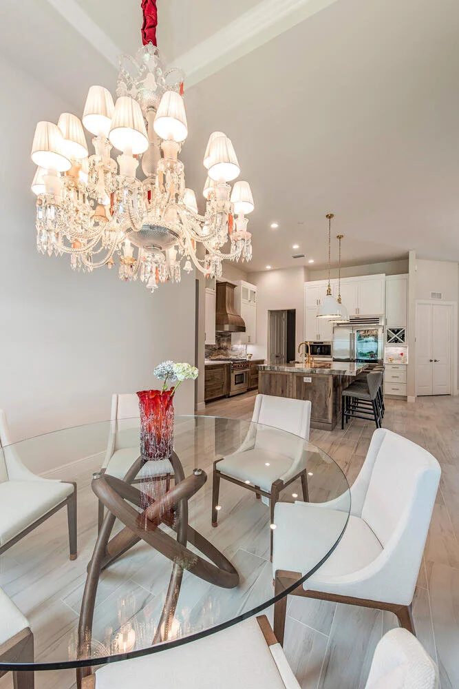 A dining area with a glass table and six white upholstered chairs, a red and black vase with white flowers, a large chandelier, and a kitchen in the background with white cabinets and a central island.