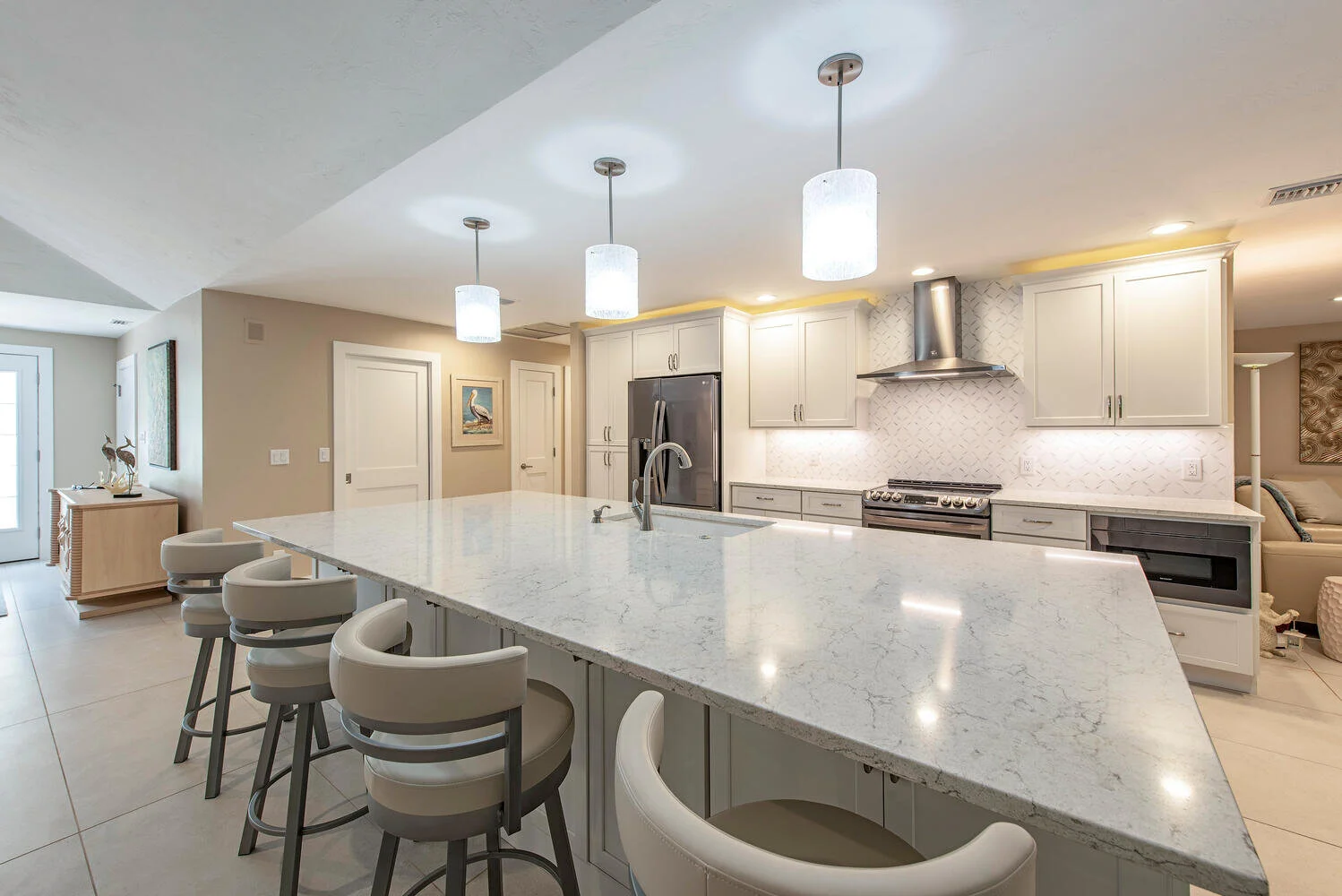 A modern kitchen with a large white marble island, pendant lights hanging above, gray cabinetry, stainless steel refrigerator, stove, and a decorative backsplash, with a small living area visible to the right.
