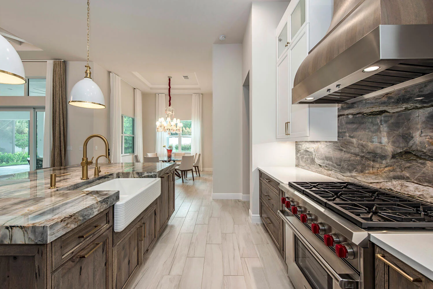 Modern kitchen with marble countertops, wooden cabinets, stainless steel stove, white sink, brass fixtures, pendant lighting, and a dining area with a chandelier and windows showing outdoor greenery.