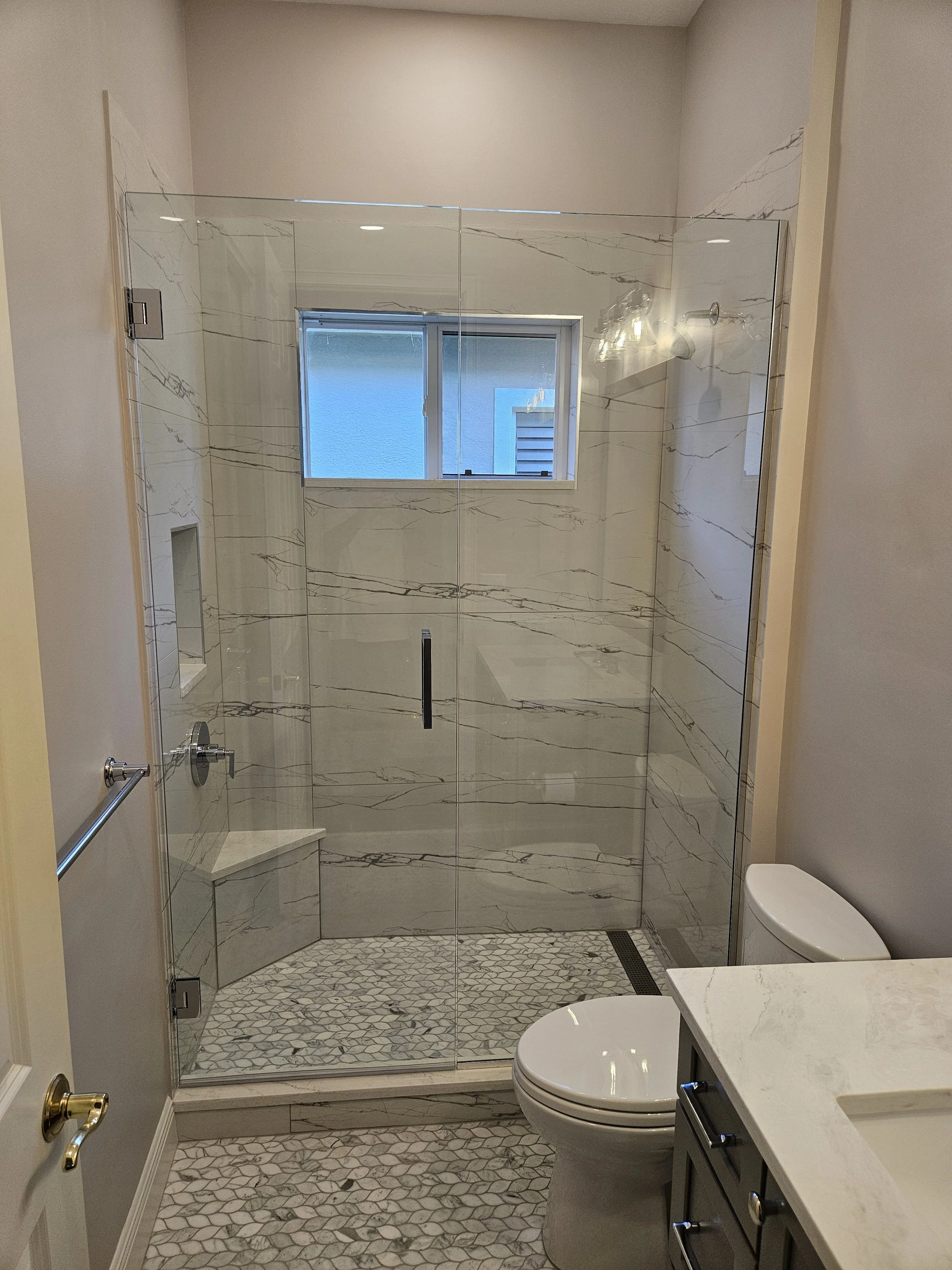 Bathroom with a glass shower enclosure, marble walls, a window, a toilet, and a vanity with a white countertop and gray cabinetry.