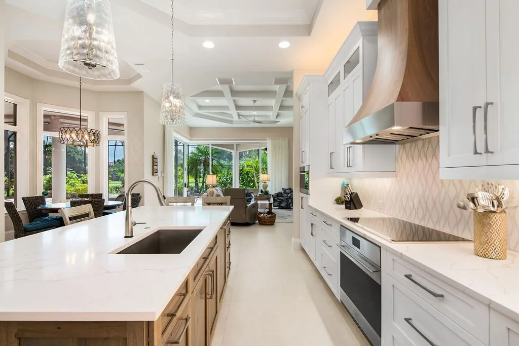 Open-concept kitchen with white cabinets, marble countertops, and a central island with a sink; view into living and dining areas with large windows and outdoor scenery.