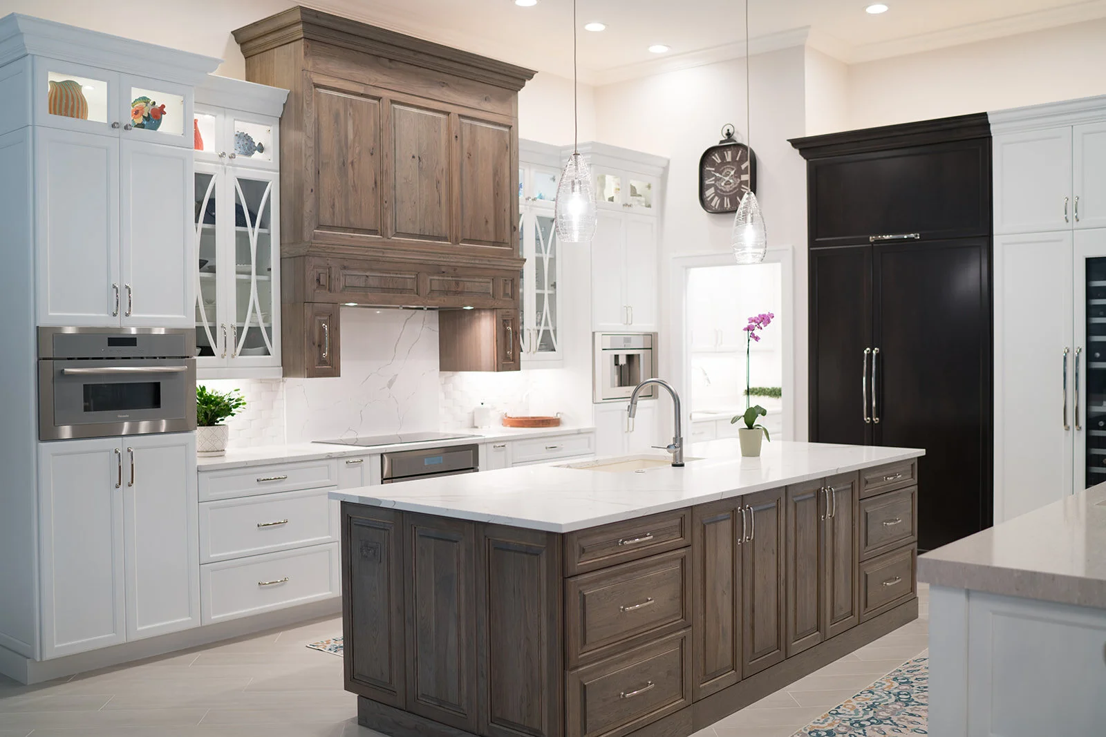 A modern kitchen with white and dark wood cabinets, a white marble backsplash, and a central island with a white countertop and dark wood cabinets. There are hanging pendant lights over the island, a potted orchid on the window sill, and a built-in m