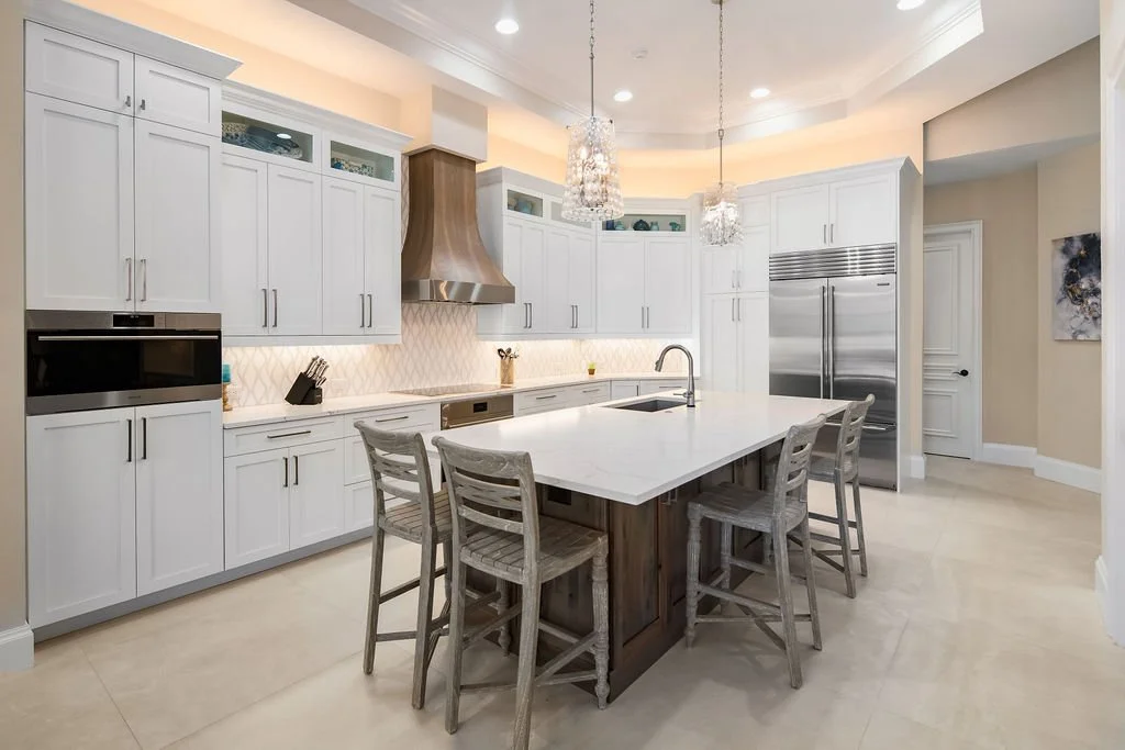 Bright modern kitchen with white cabinetry, a large island with a white marble top, four wooden chairs, stainless steel appliances including a refrigerator and oven, a wooden range hood, and pendant lights.