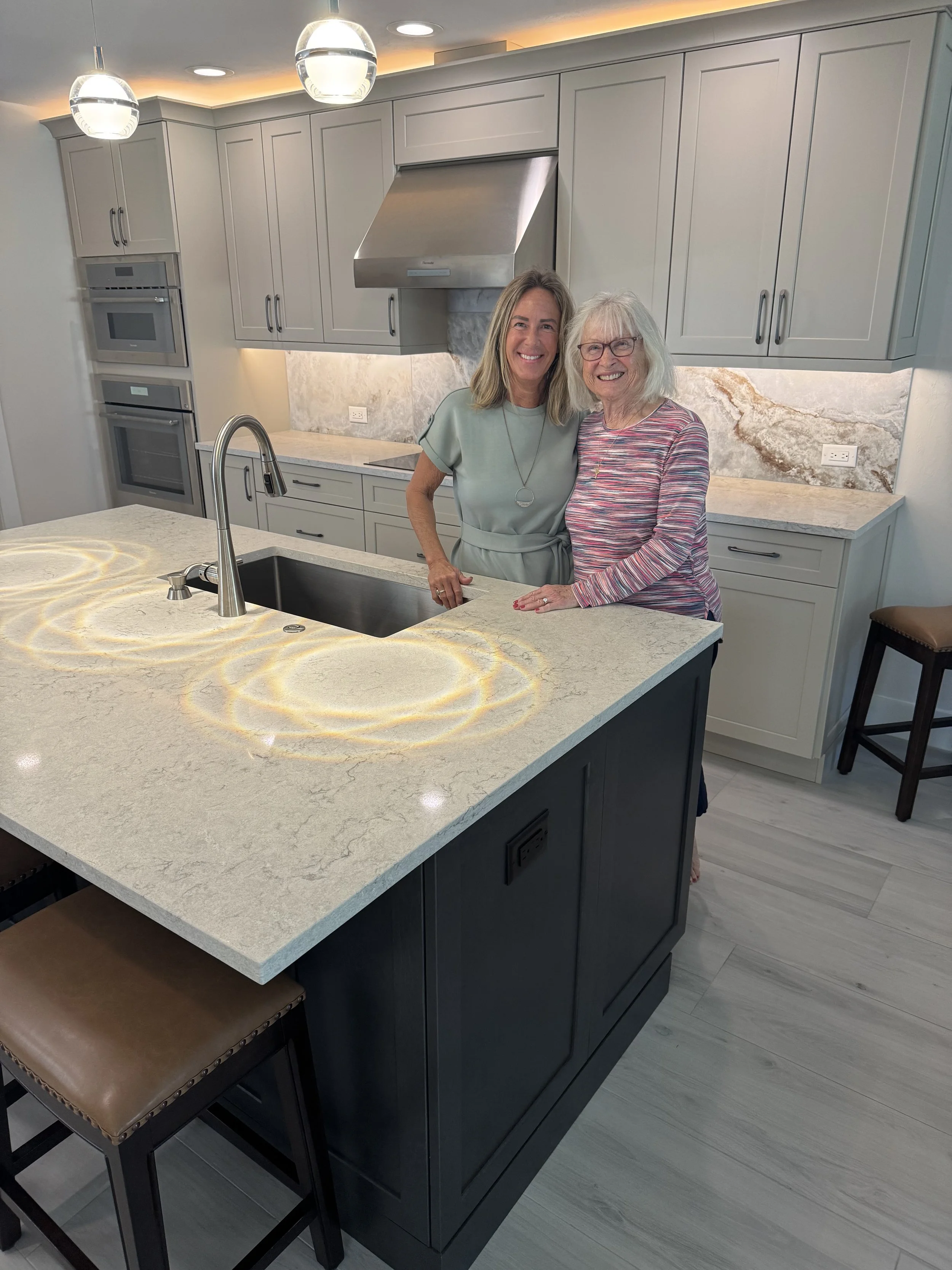 Two women smiling in a modern kitchen, standing behind a kitchen island with a marble countertop. One woman has light brown hair and wears a light green dress. The other has white hair, wears glasses, and a multicolored striped top. The kitchen featu