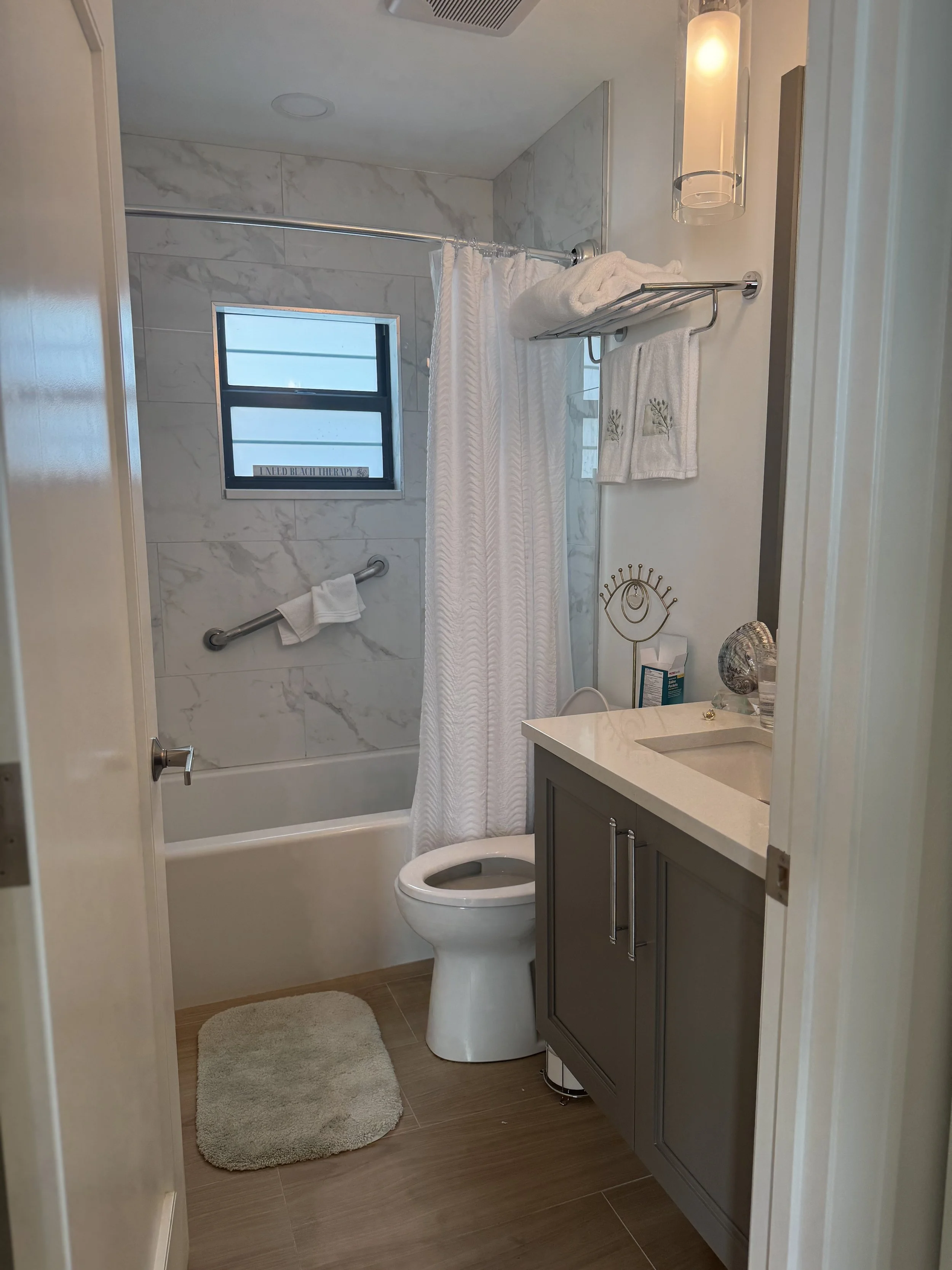 A modern bathroom with a shower area, a window, a toilet, and a sink vanity with a gray cabinet. The shower has a marble wall and a curtain, with a towel hanging on a rail. There is a small rug in front of the toilet, decorative items on the sink, an