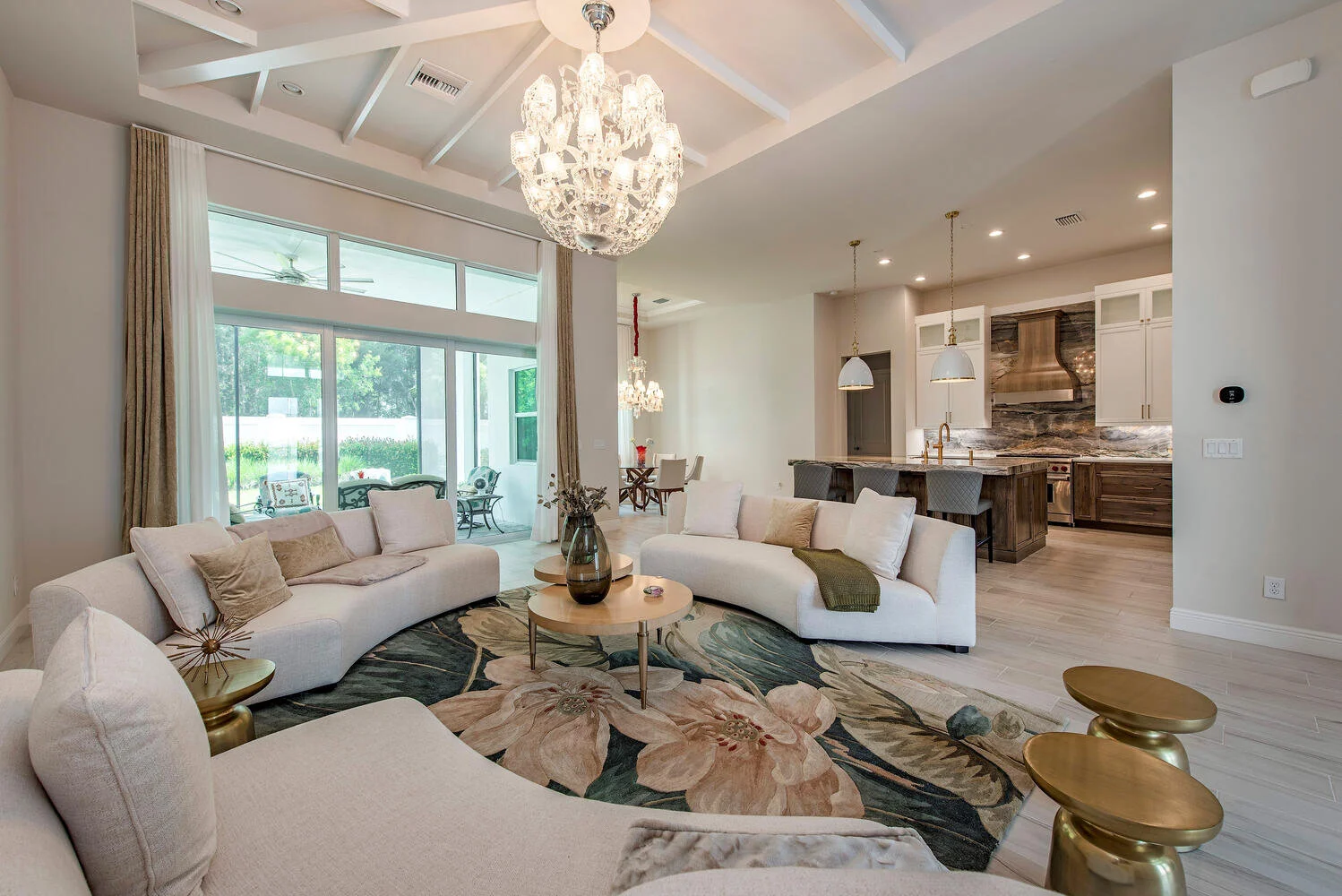 Living room with white sofas, floral area rug, large glass sliding doors leading outside, chandelier, and open kitchen with wooden cabinetry and white pendant lights.