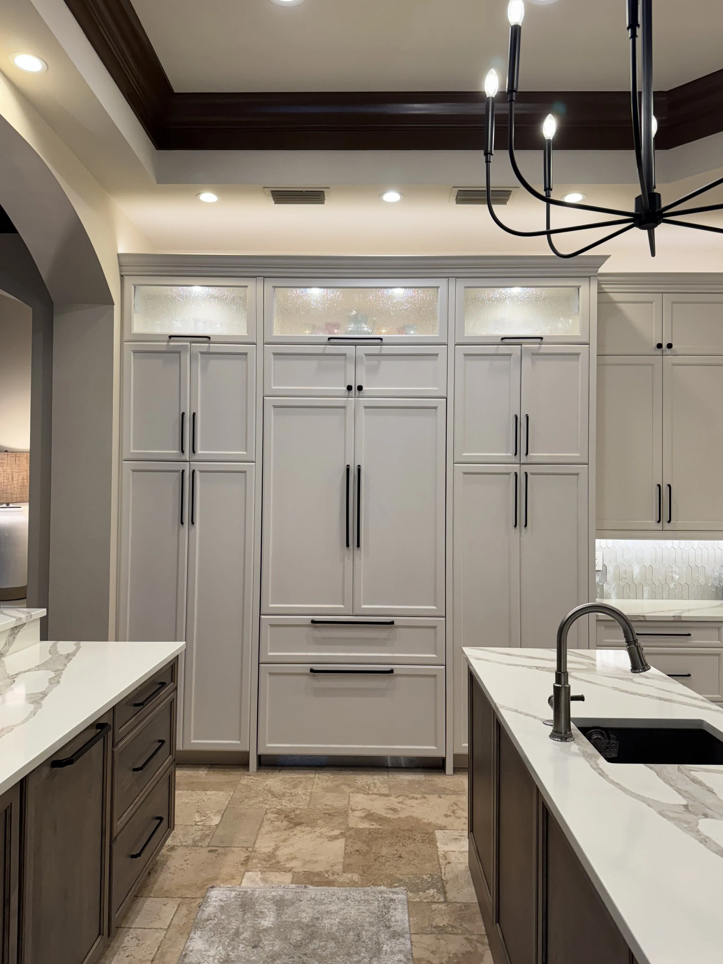 Modern kitchen with white cabinets with black handles, a kitchen island with a marble countertop, stainless steel faucet, and beige tiled floor. Recessed lighting and a black chandelier hanging from the ceiling.