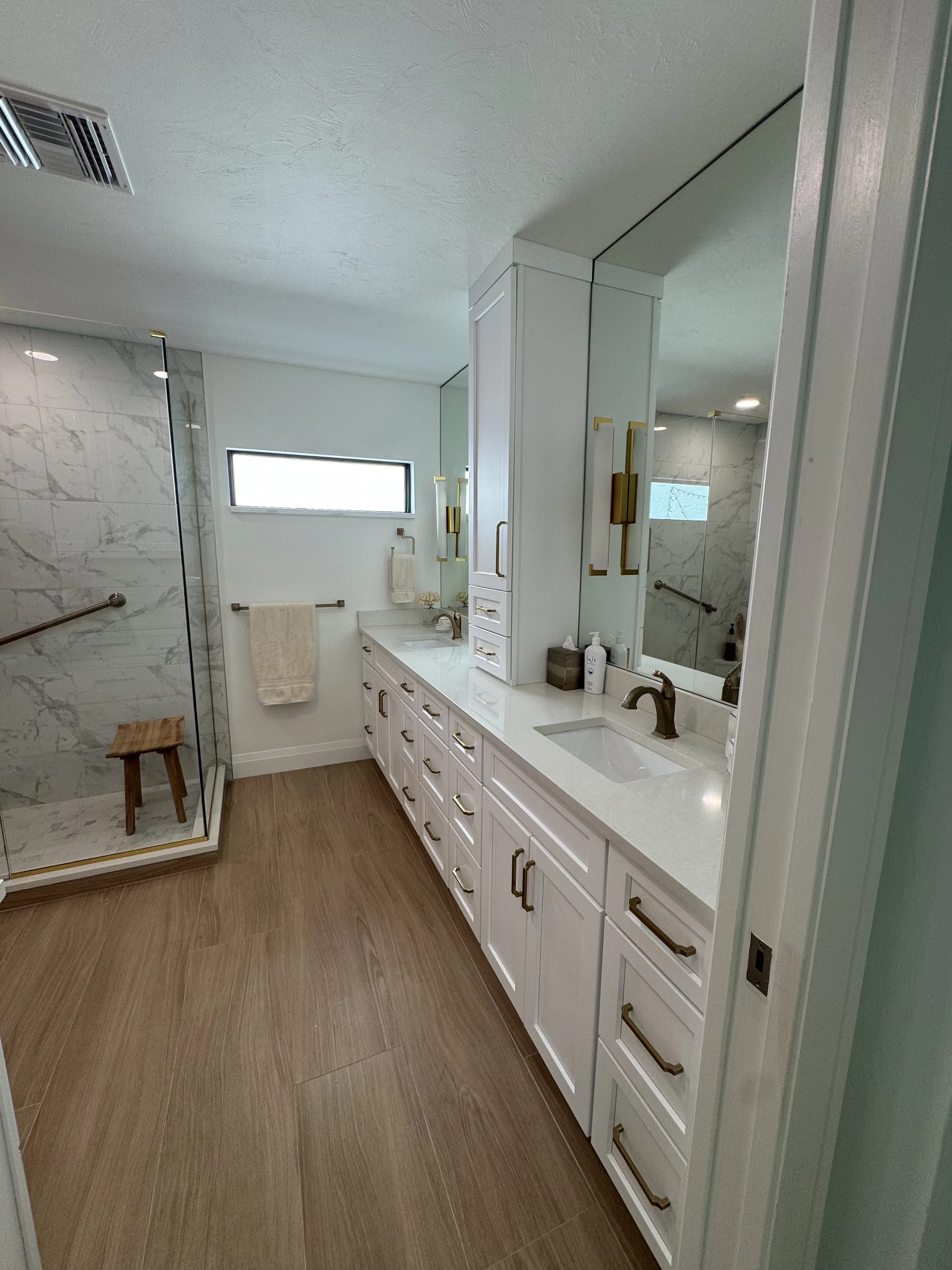 A modern bathroom with a double vanity, white cabinets with gold handles, and a marble shower enclosure with a wooden stool inside. The bathroom has wood-look tile flooring, a small horizontal window, and a mirror above the sinks. There are also towe