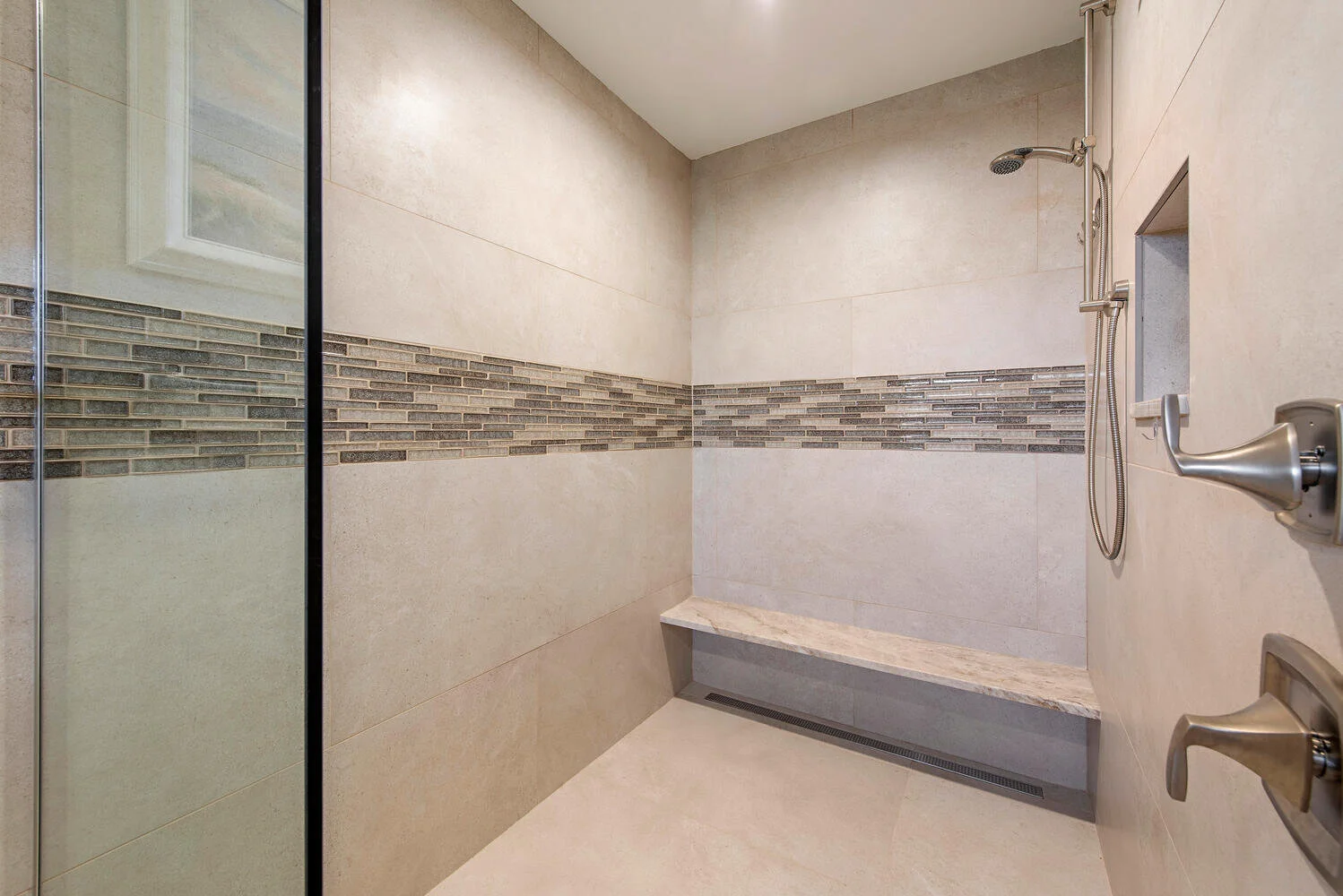 Shower area with beige tile walls, a horizontal strip of decorative tile, a marble bench, rain showerhead, hand-held shower, and a glass door.