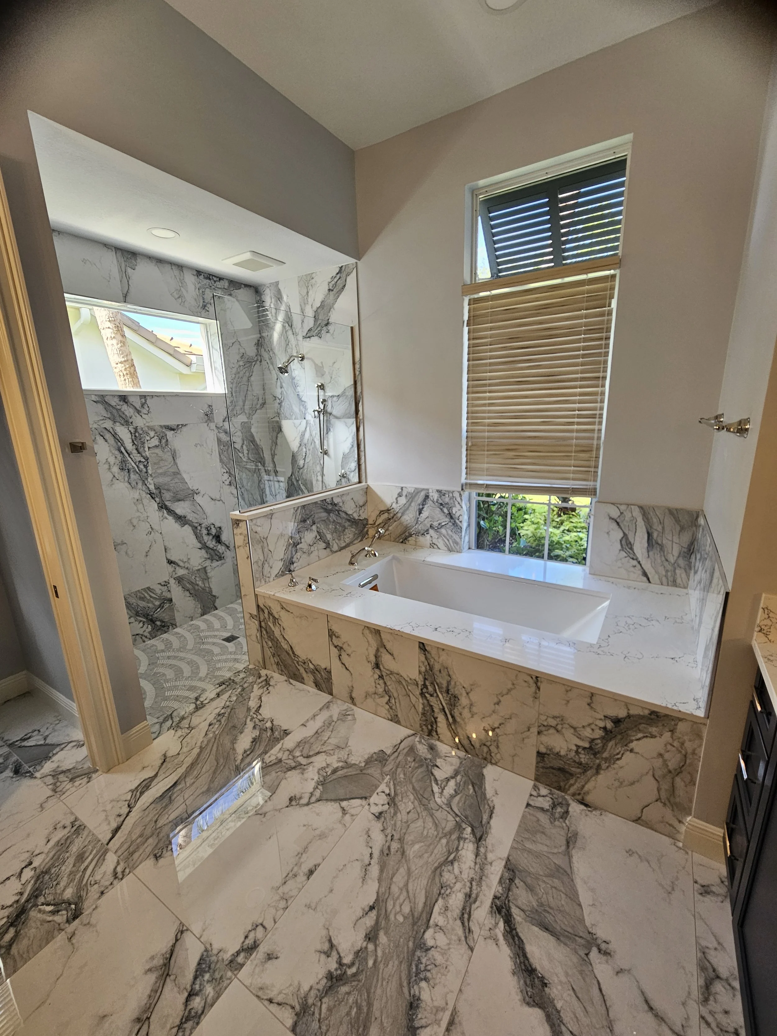 Bathroom with marble walls and floors, a bathtub, shower area, window with blinds, and a toilet.