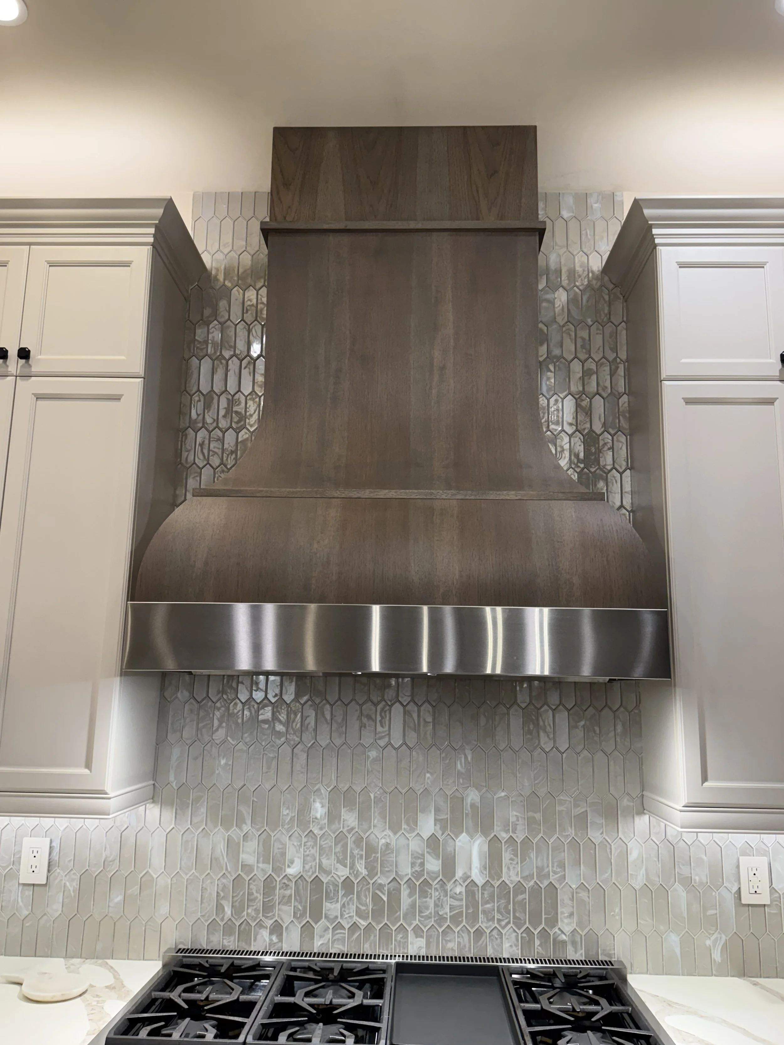 Photo of a modern kitchen stove with a gas cooktop, silver range hood, decorative tile backsplash, and white cabinetry.