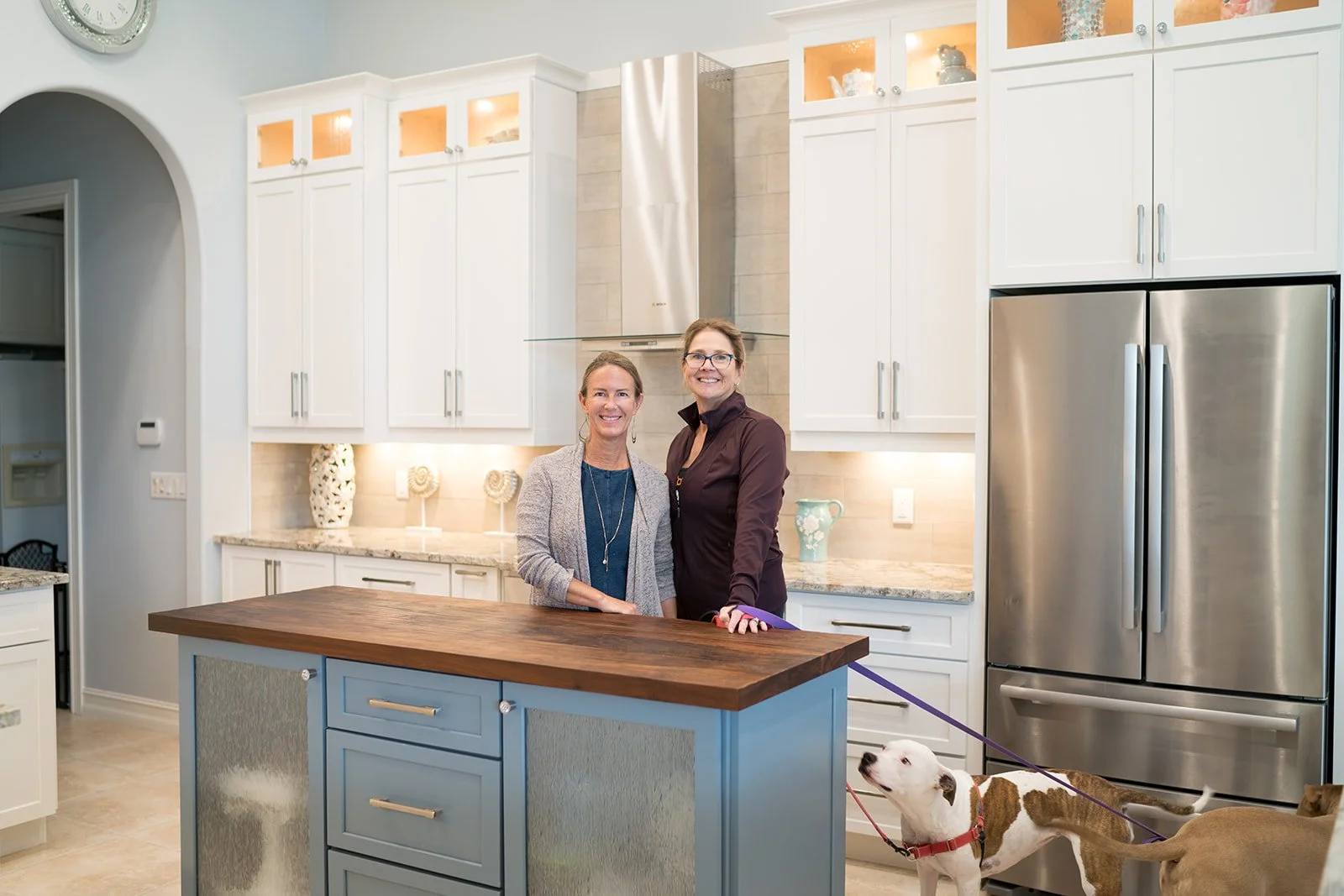 Two women standing in a modern kitchen with white cabinets and stainless steel appliances, smiling at the camera, with a dog on a leash.