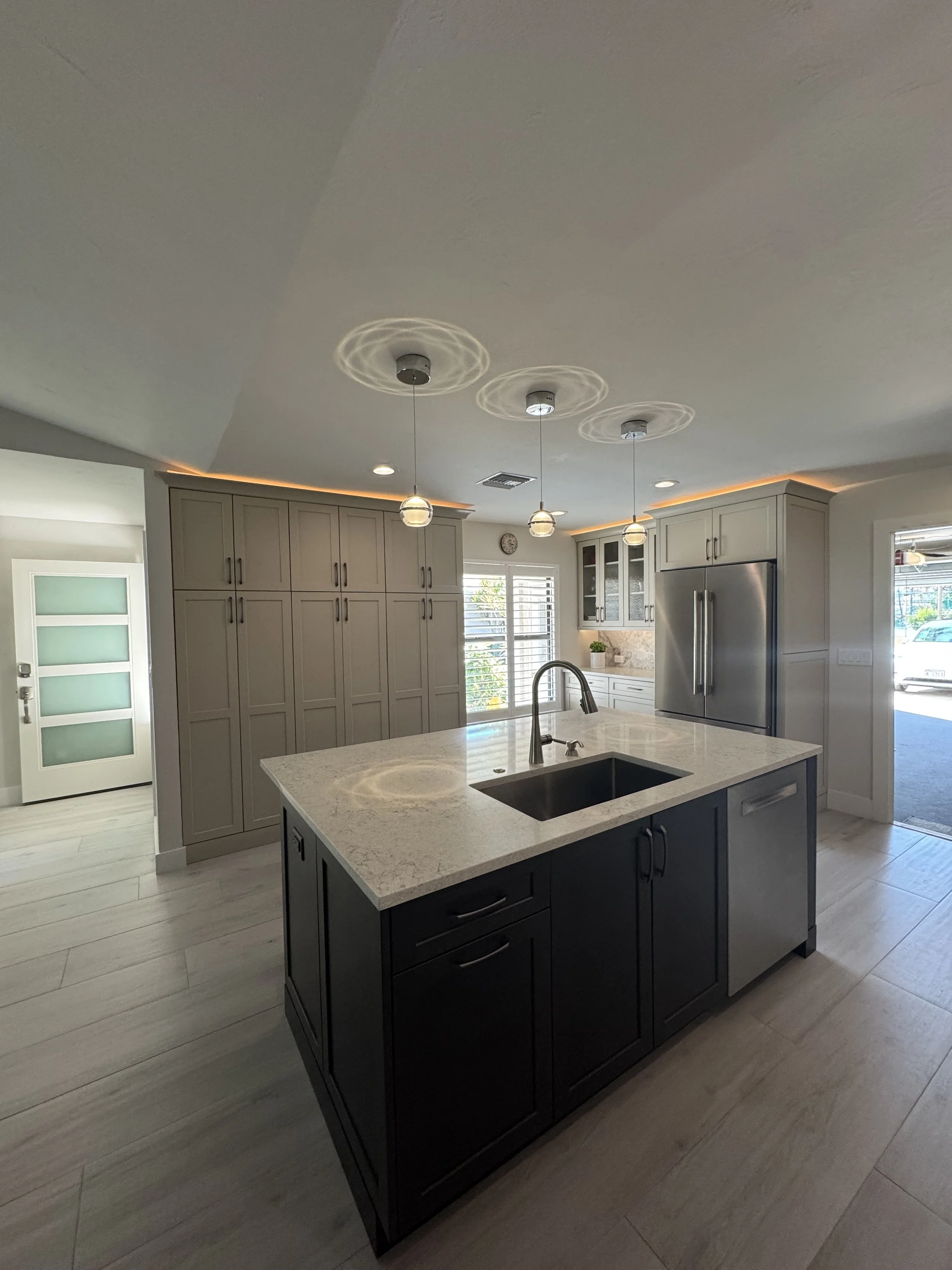 Modern kitchen with white cabinetry, stainless steel refrigerator, granite island with sink, pendant lights, and natural light from windows.