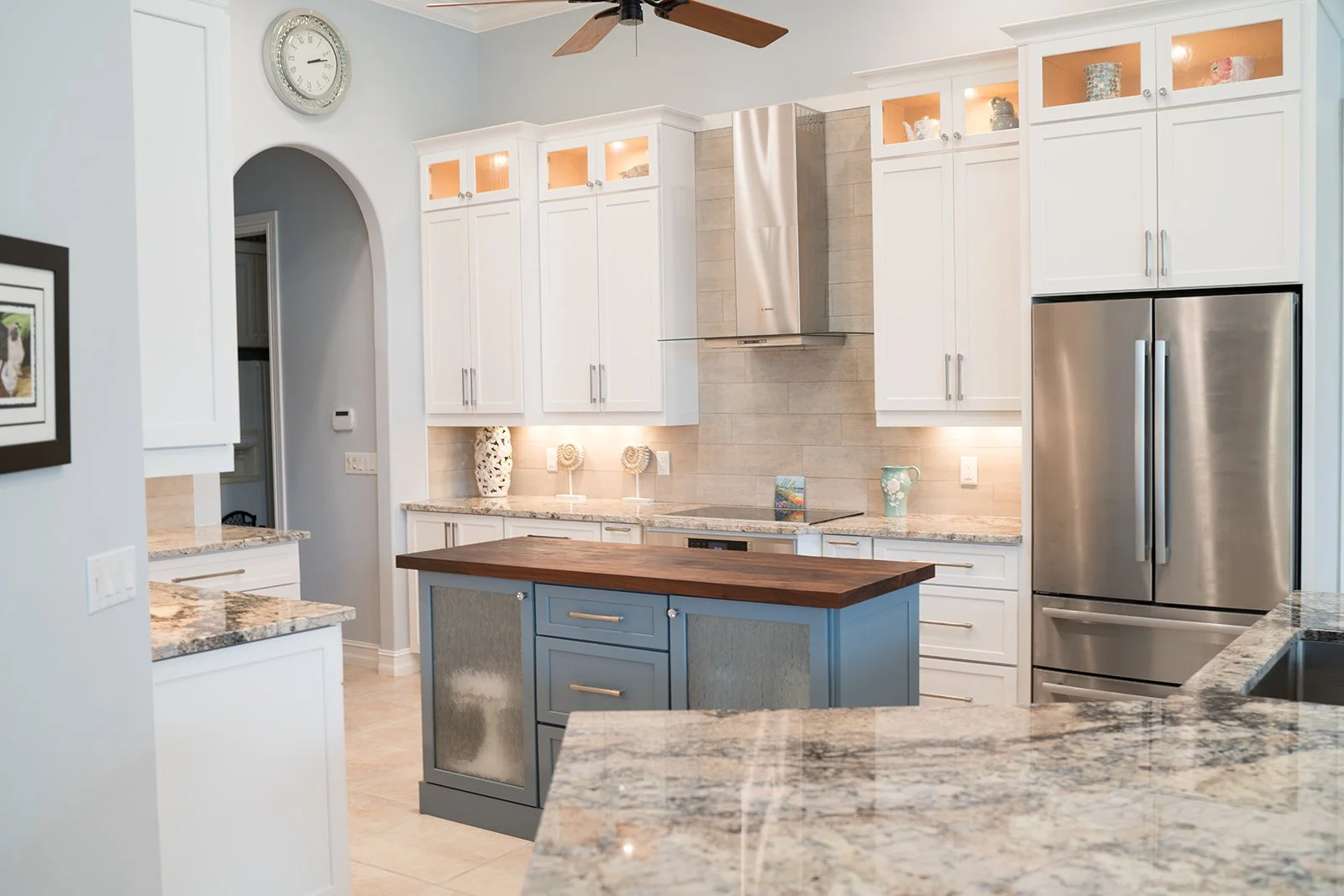 Modern kitchen with white cabinets, a stainless steel refrigerator, a central island with a wooden countertop, and decorative vases on the counters.