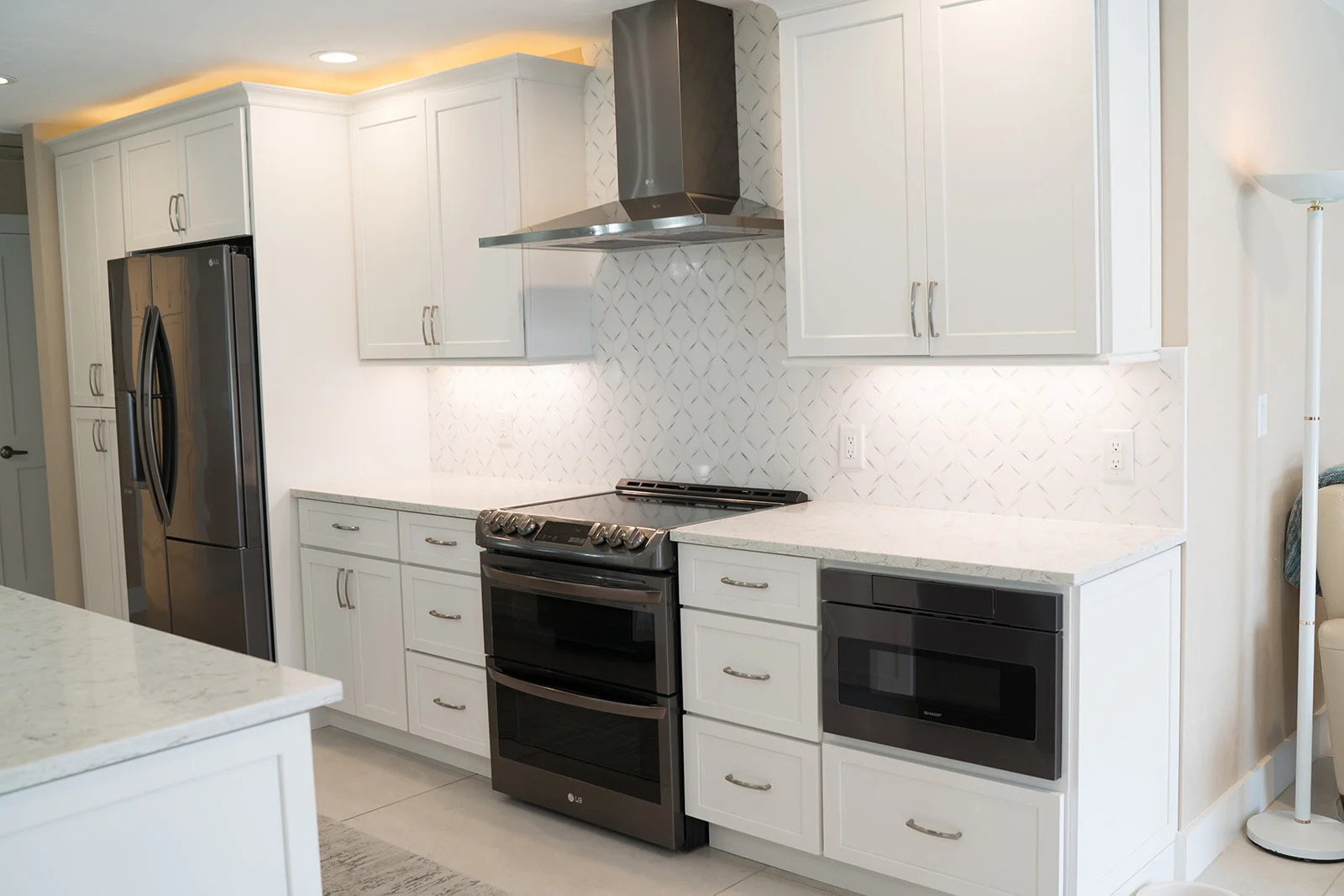 Modern kitchen with white cabinets, stainless steel refrigerator, black oven and microwave, white quartz countertops, patterned white backsplash, and a stainless steel range hood.