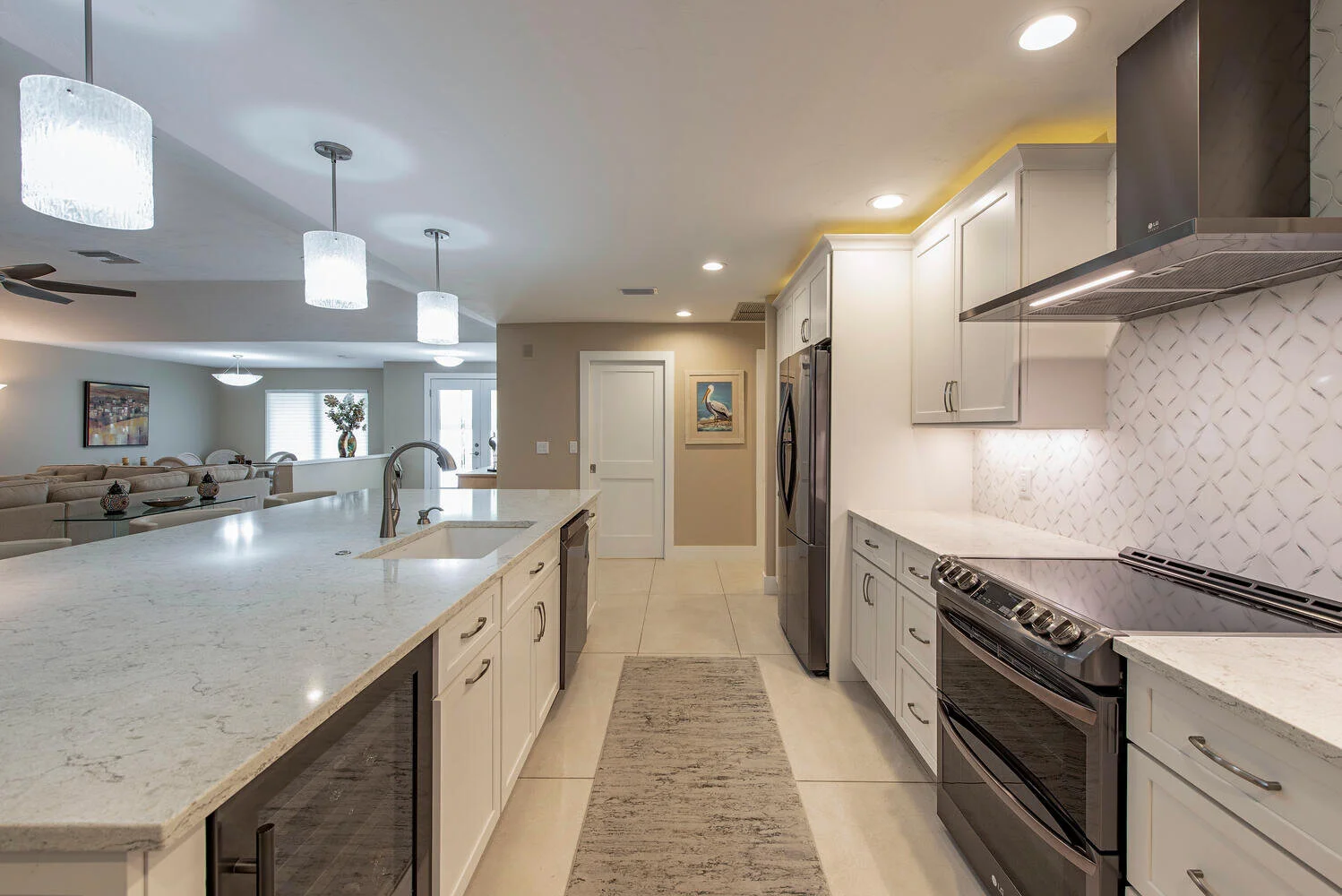 Modern kitchen with white cabinets, stainless steel appliances, marble backsplash, and a large island with a double sink and bar seating. Open concept living space in background.