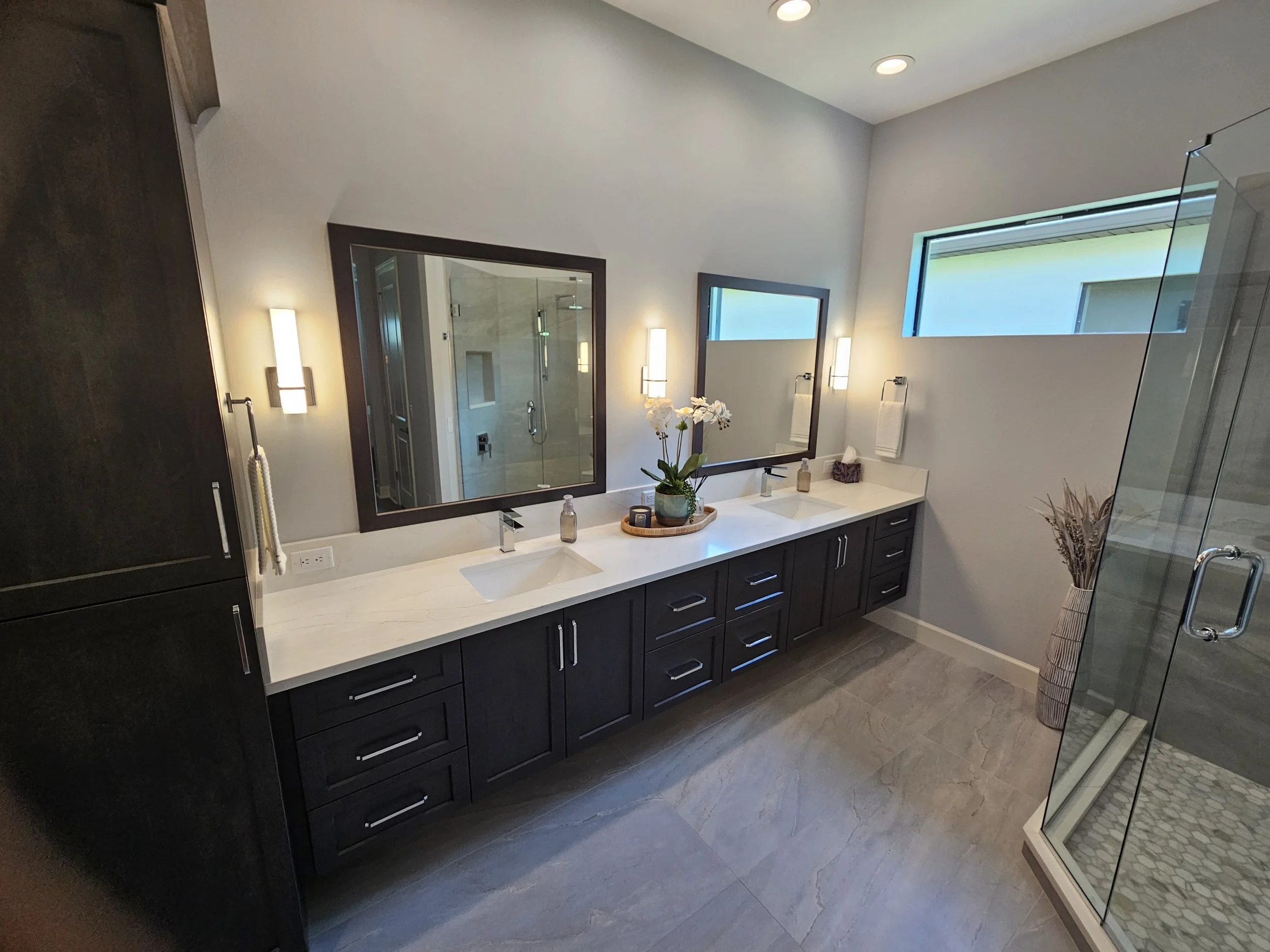 Modern bathroom with a wide countertop featuring two sinks, two large mirrors with dark frames, and a potted orchid plant. There are two vertical light fixtures on the wall, a window above, and a glass shower enclosure to the right.