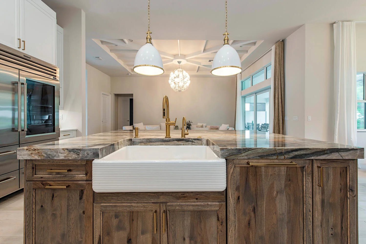 A modern kitchen with a marble island, a white farmhouse sink, brass faucet, hanging pendant lights, and a view into a dining area with a chandelier and large windows.