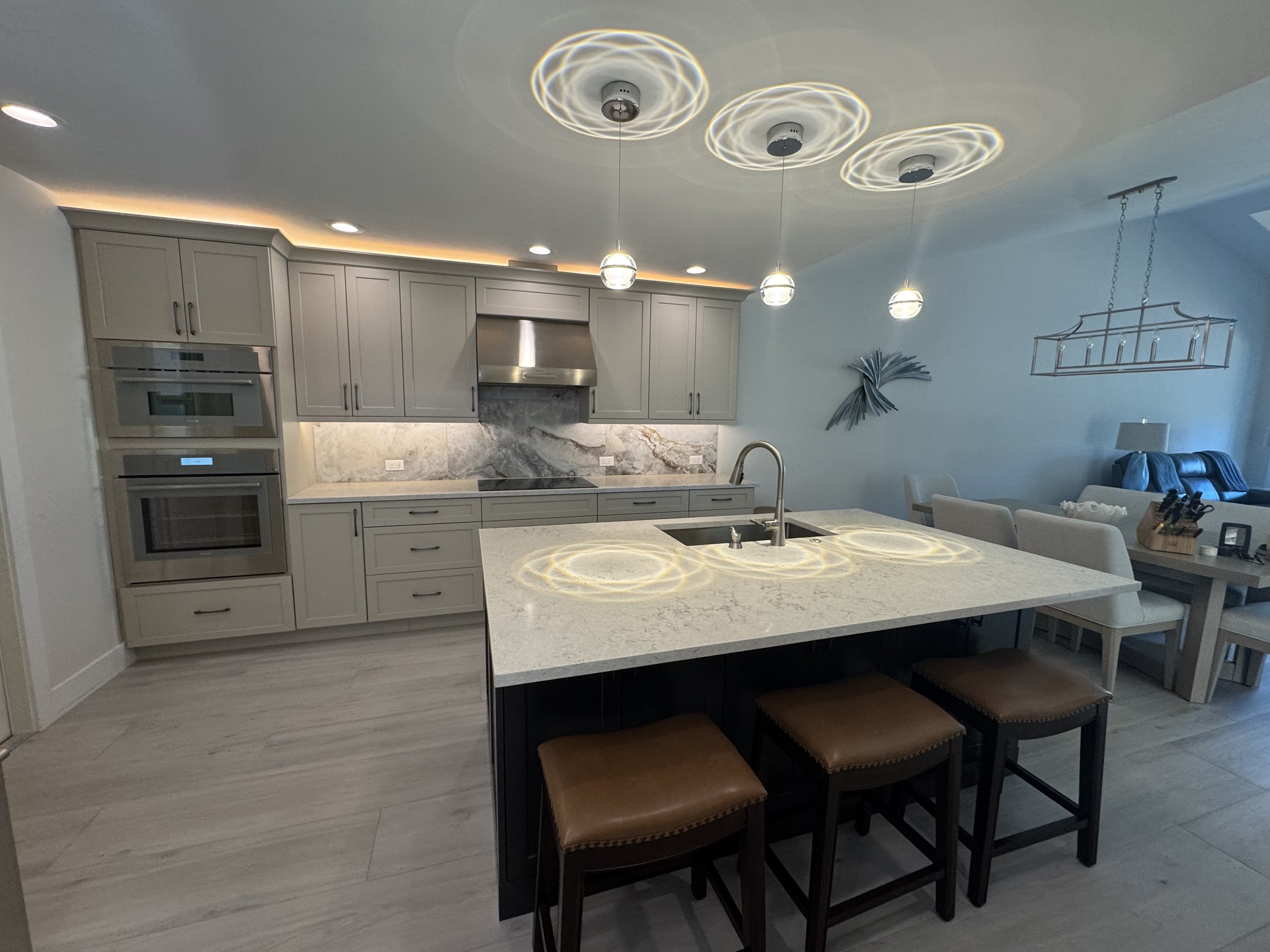 Modern kitchen with white cabinets, marble backsplash, and white countertop island. Three pendant lights with circular patterns are hanging from the ceiling, casting swirling light reflections on the ceiling. The kitchen has built-in appliances and a