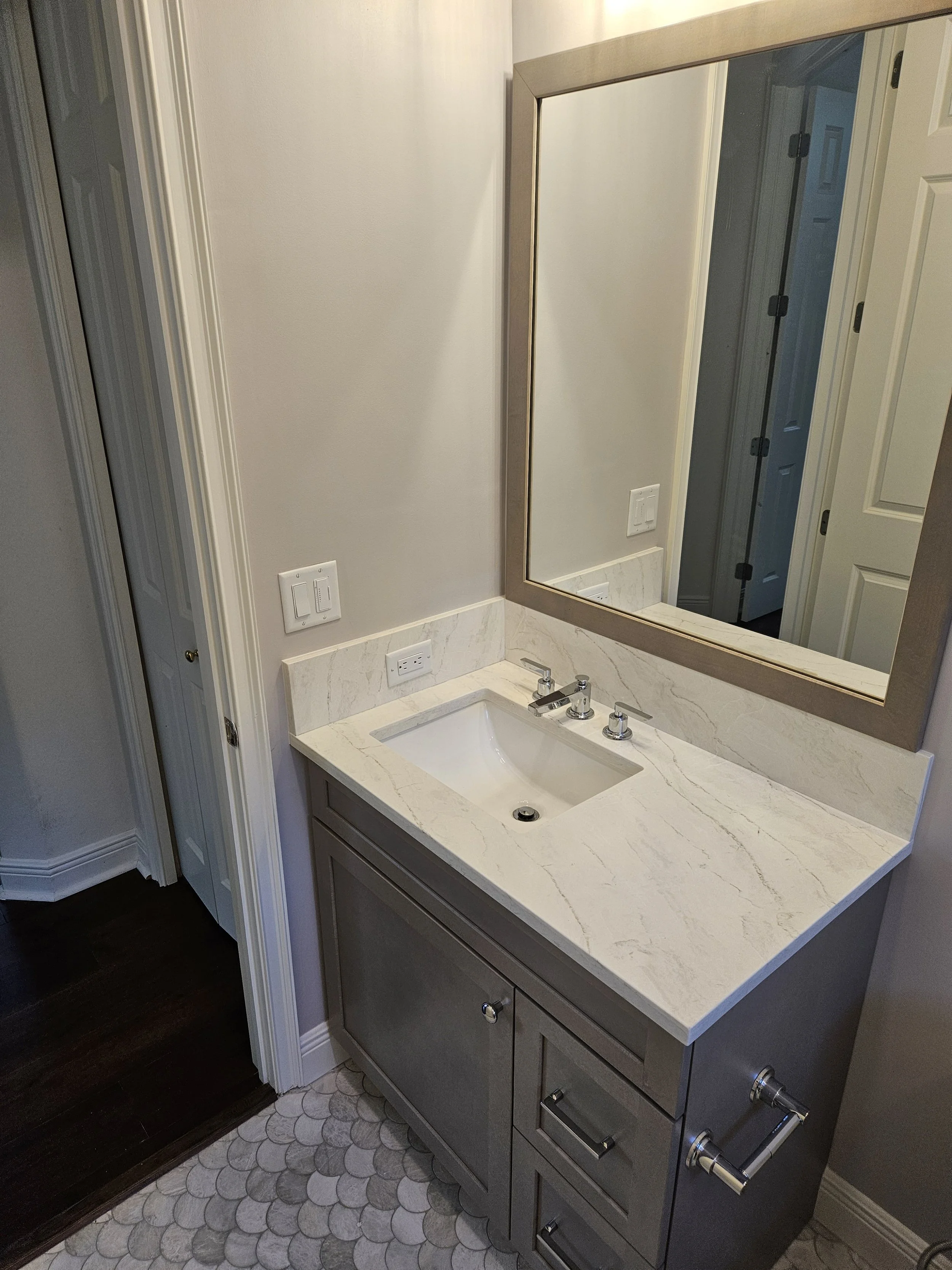 Bathroom with a mirror, beige countertop with a sink, gray cabinet with multiple drawers, wooden floor, white walls, and a door in the reflection.