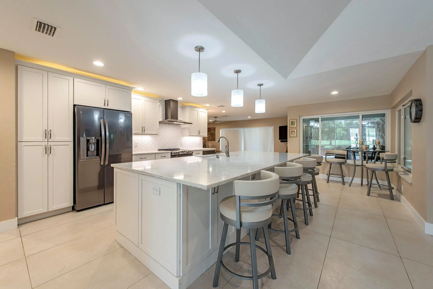 Modern kitchen with white cabinets, stainless steel refrigerator, large marble island with bar stools, pendant lighting, and a dining area near sliding glass doors.