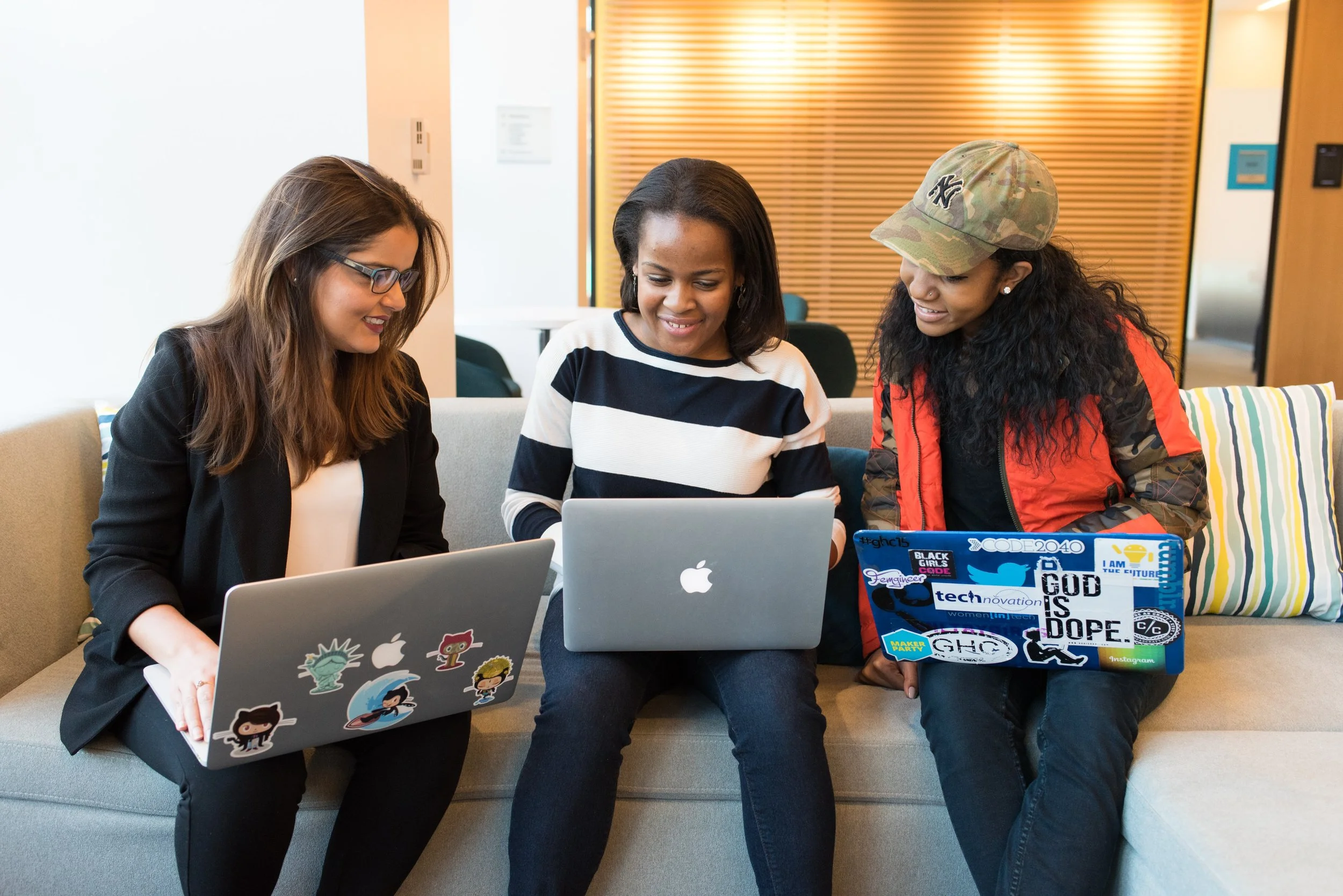 Three women sitting on a beige couch, using laptops, in a modern office setting.