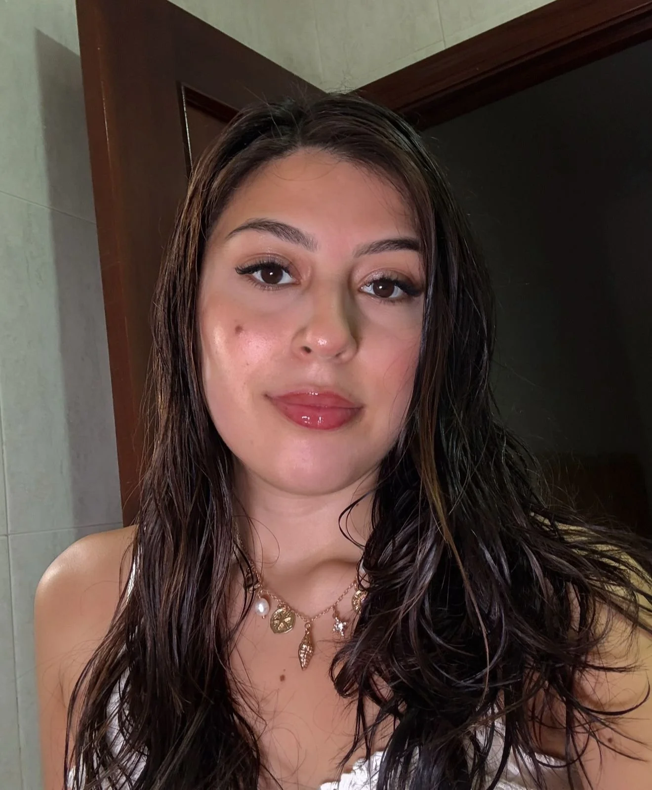 A young woman with long, dark, wavy hair and makeup, wearing layered necklaces, standing indoors, with a door and tiled wall in the background.