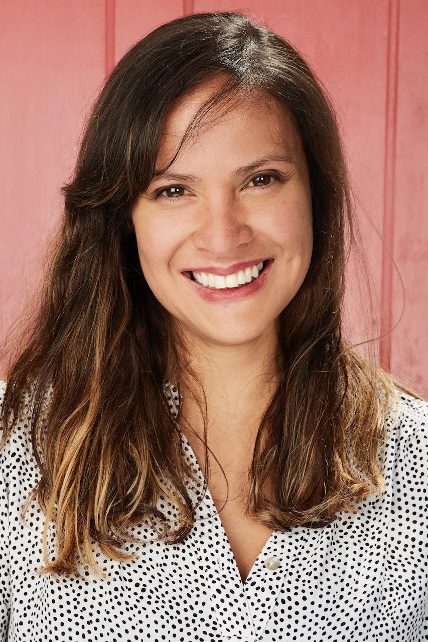 A smiling woman with long brown hair wearing a white blouse with black polka dots, standing in front of a pink wooden background.