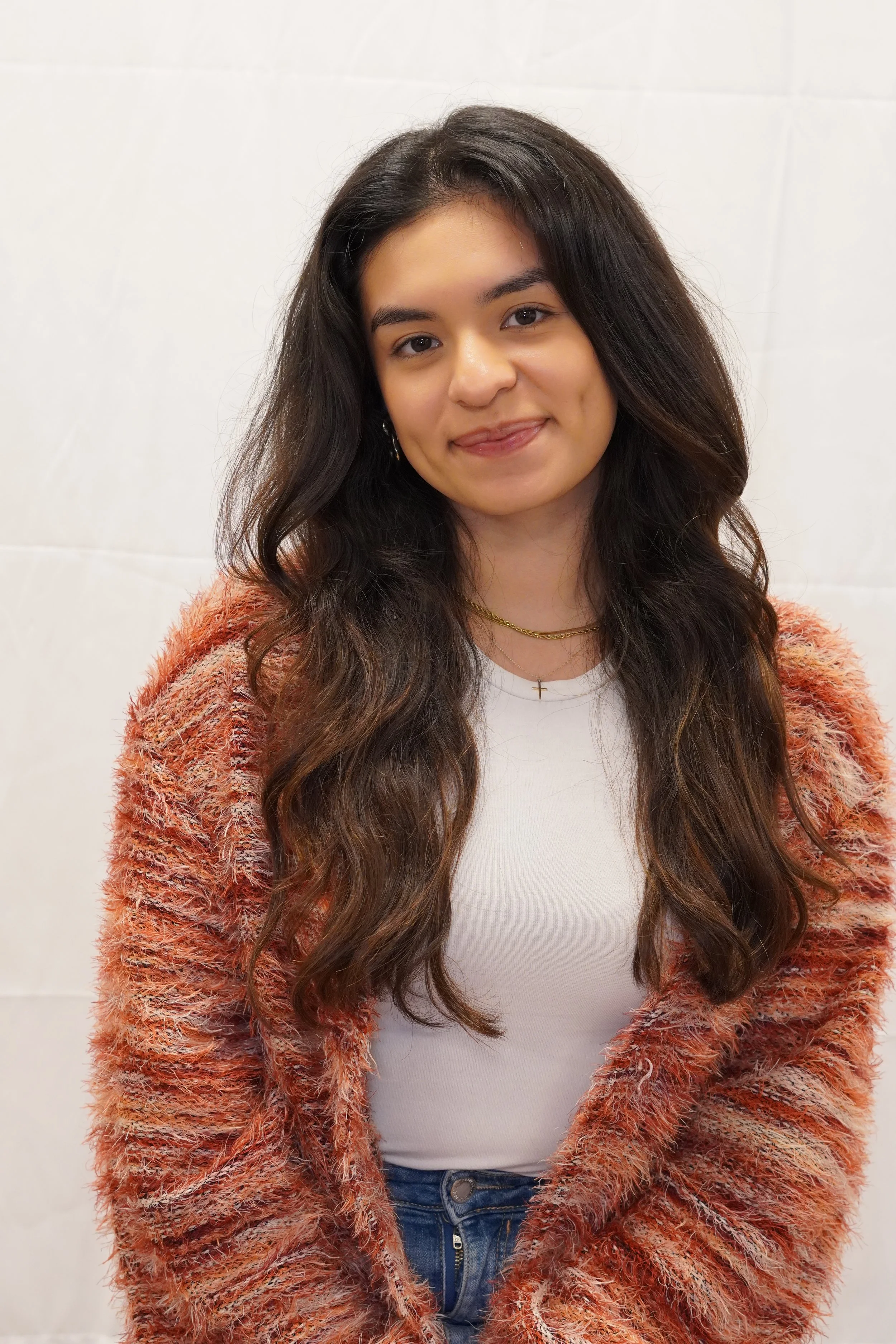 A young woman with long, wavy brown hair smiling at the camera, sitting on a black stool against a plain white background, wearing a fuzzy orange and pink sweater and blue jeans.