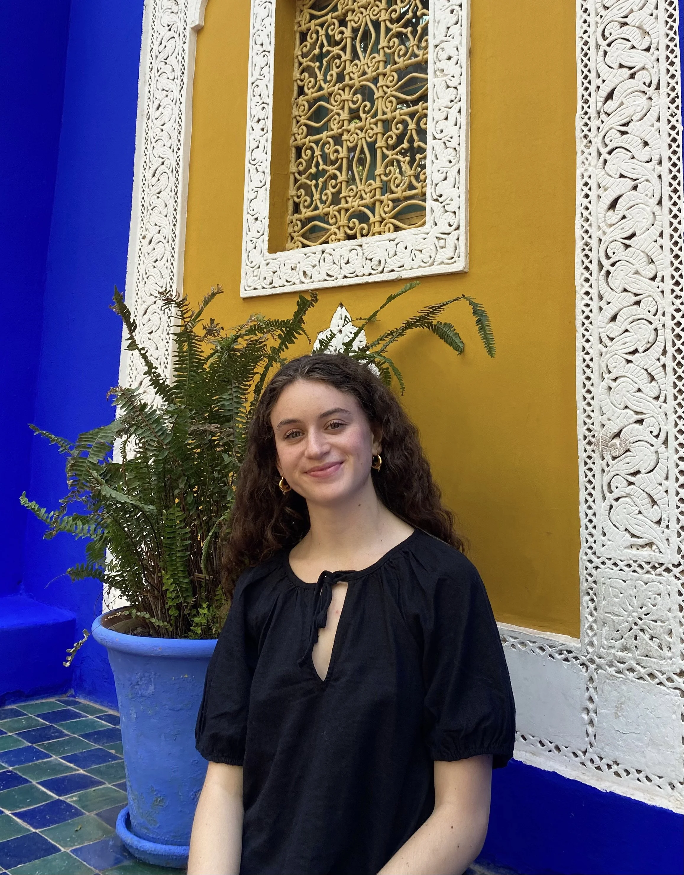 A young woman with curly hair smiling, wearing a black blouse, sitting in front of a vibrant yellow wall with ornate white carvings and a window with a decorative grille, next to a large potted fern plant against a blue wall.