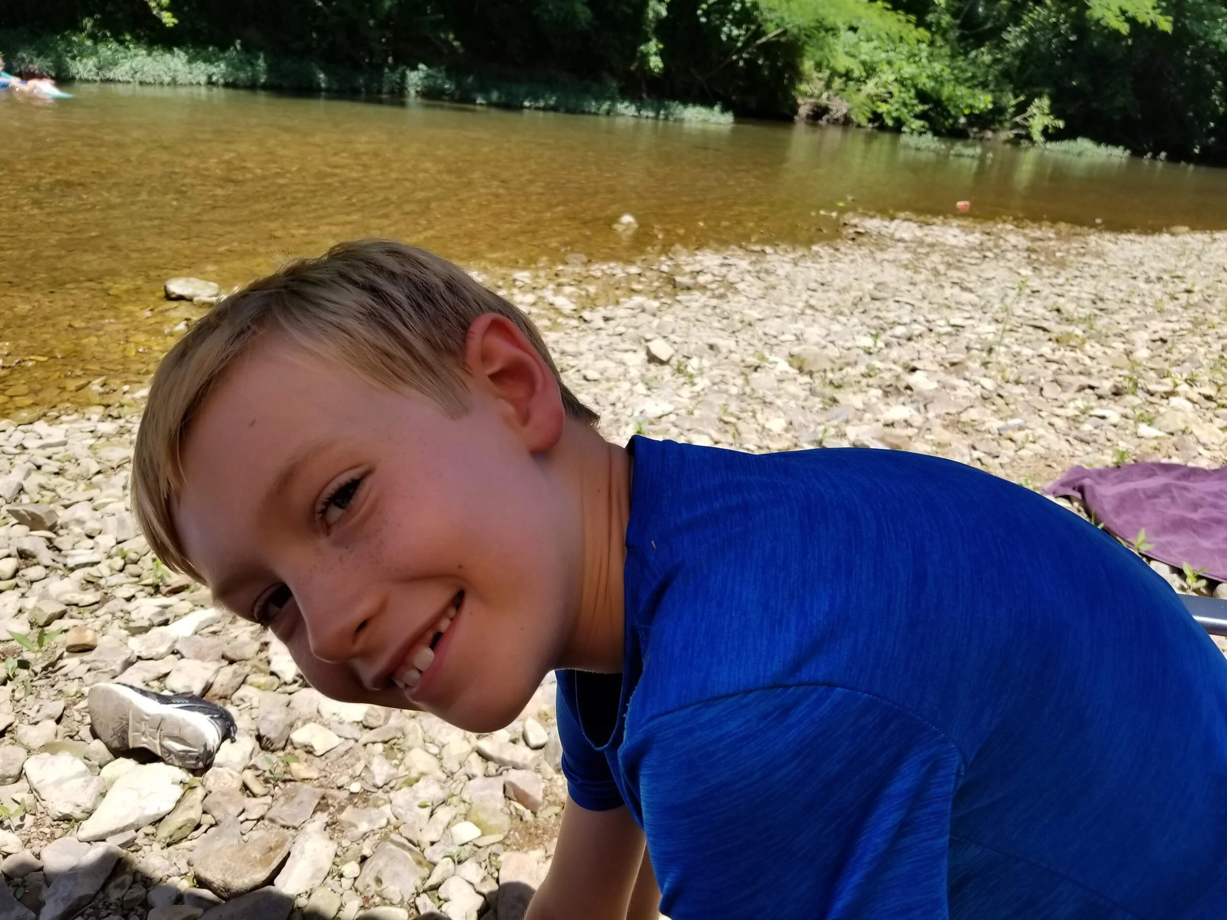 Smiling child in blue shirt near a rocky riverbank.