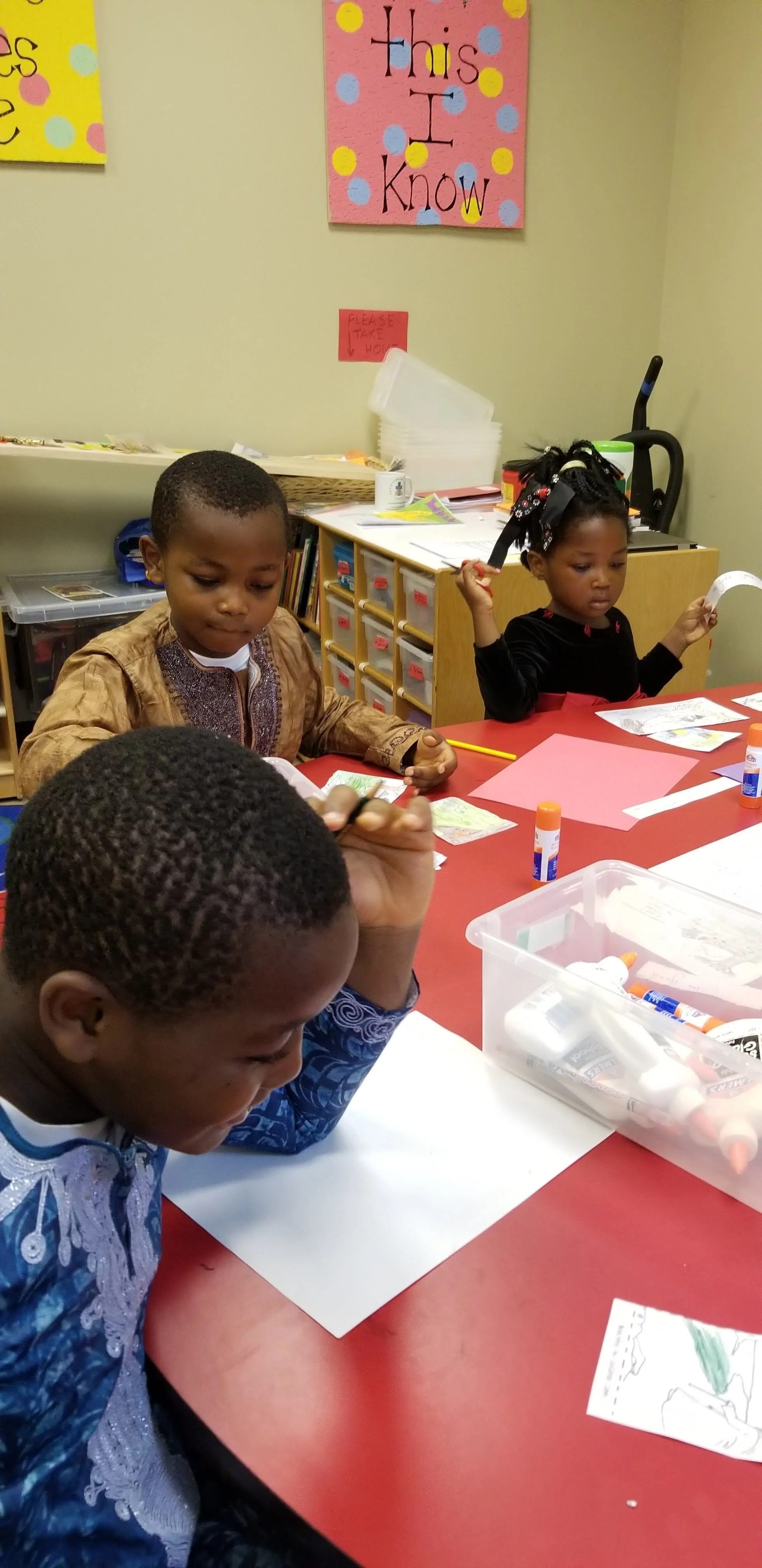 Three children at a classroom table working on arts and crafts, with supplies like paper and glue sticks on the table. A colorful "This I Know" poster is on the wall behind them.