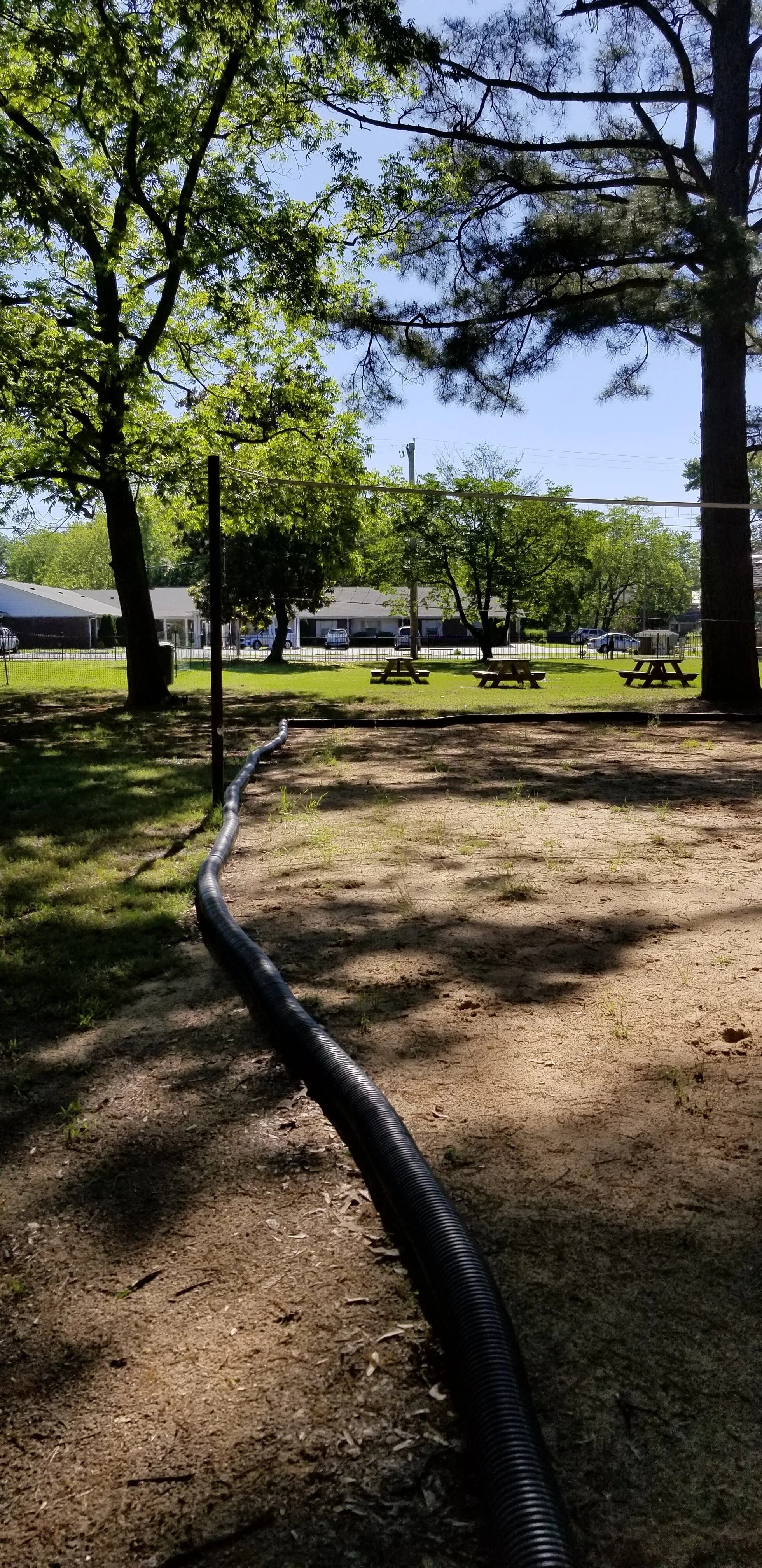 Outdoor area with trees, a volleyball net, picnic tables, and a flexible drainage pipe on the ground.