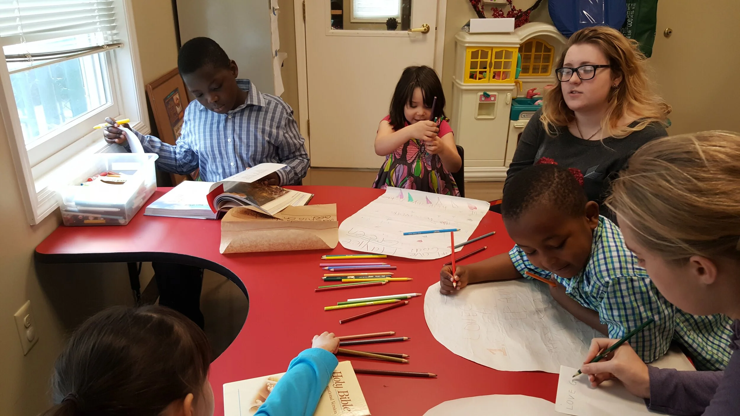Group of children and an adult sitting at a red table engaged in creative activities with pencils and paper inside a classroom.